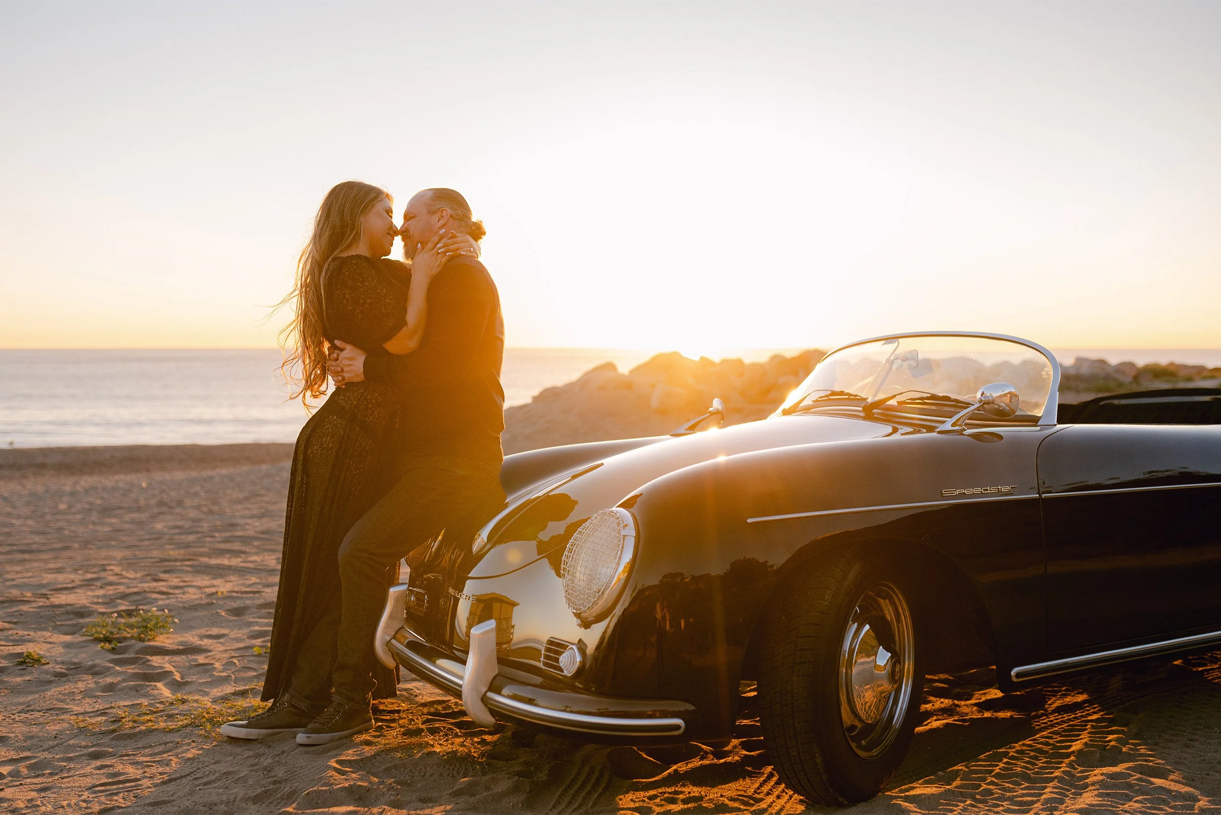 Award-winning engagement photographer in Malibu captures couple embracing beside a vintage Porsche Speedster on the beach at sunset