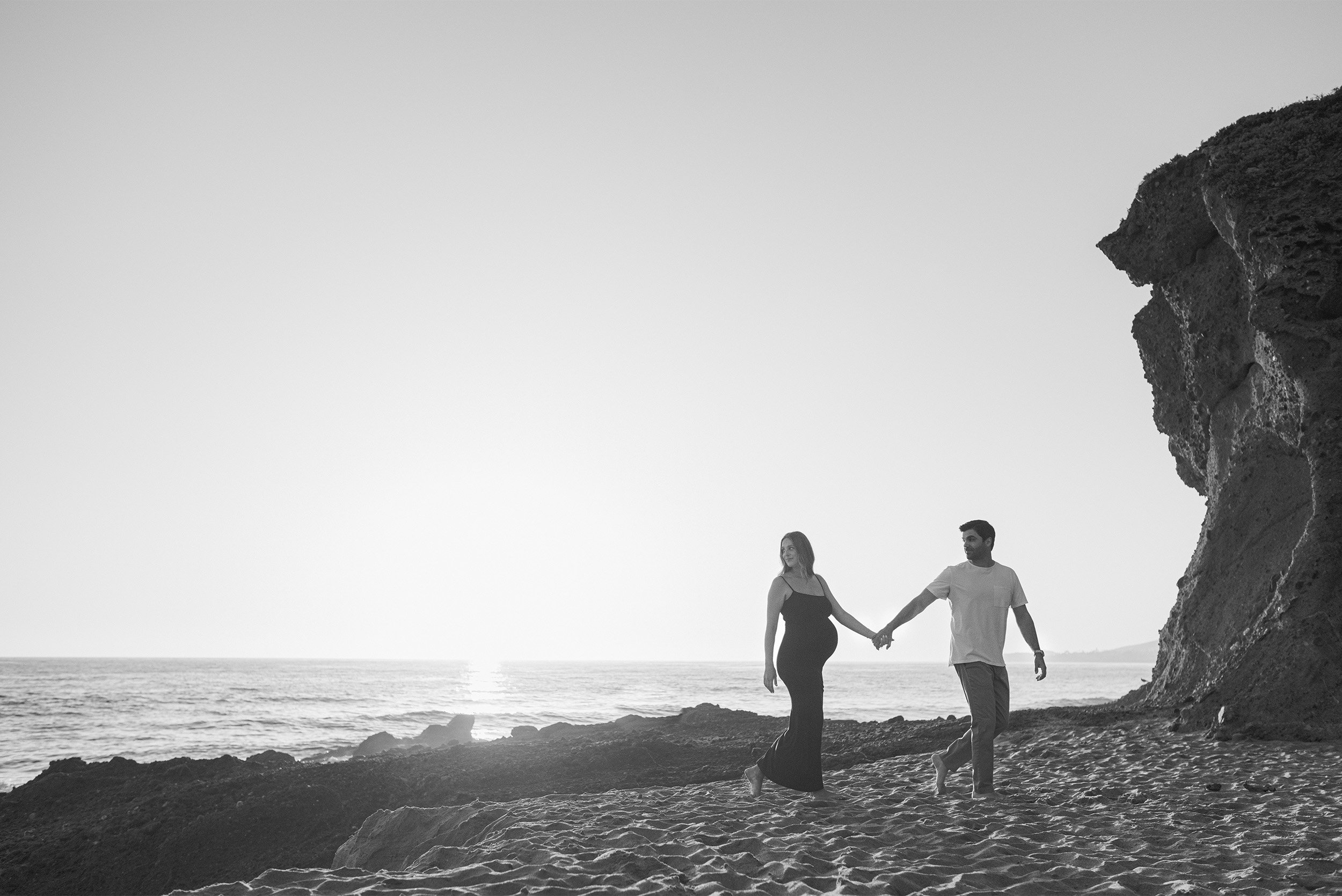 Black and white photo of pregnant couple walking along rocky shoreline, award-winning maternity photographer in Orange County