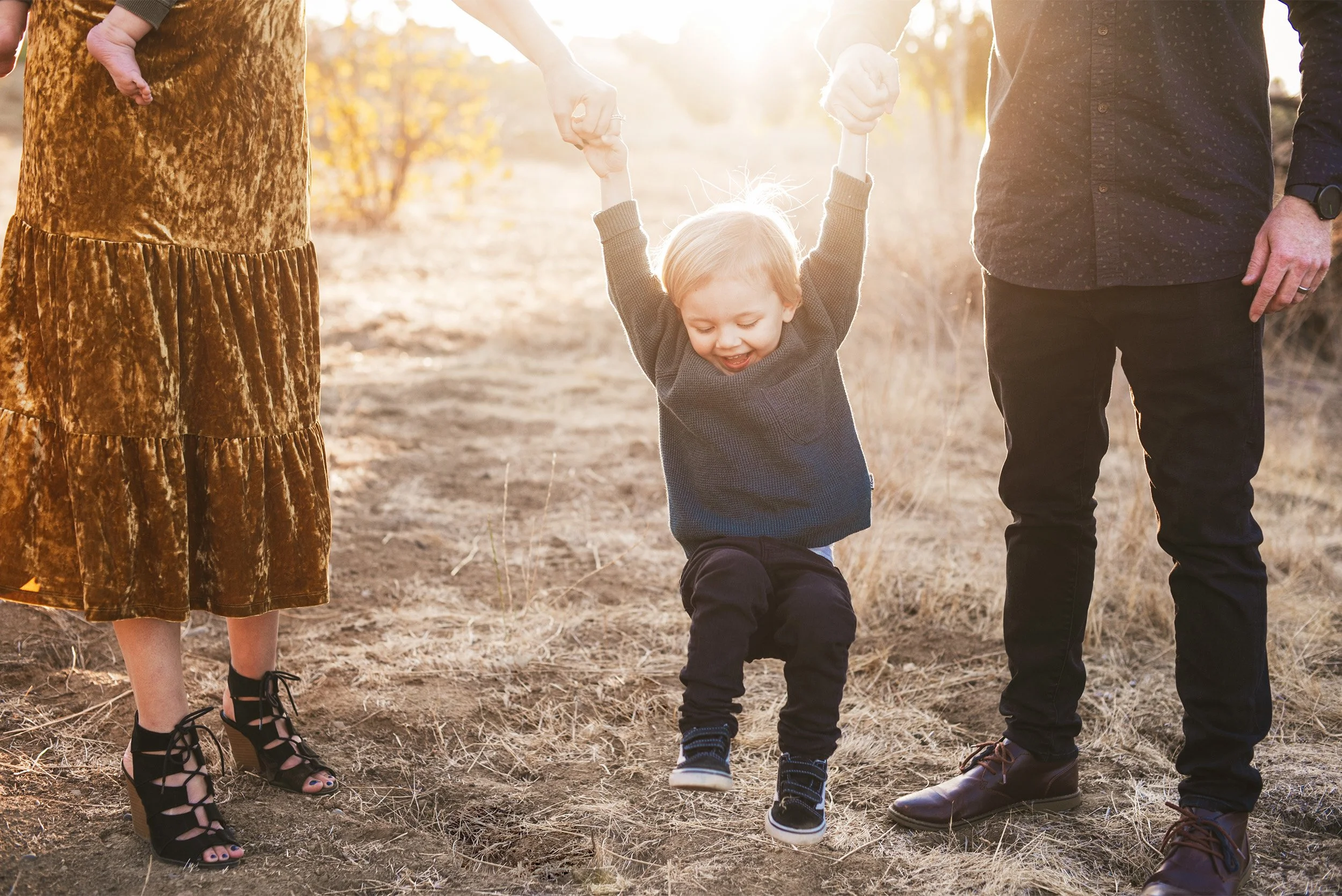 Family photographer in Orange County photographs toddler boy swinging between parents' hands in golden field at sunse