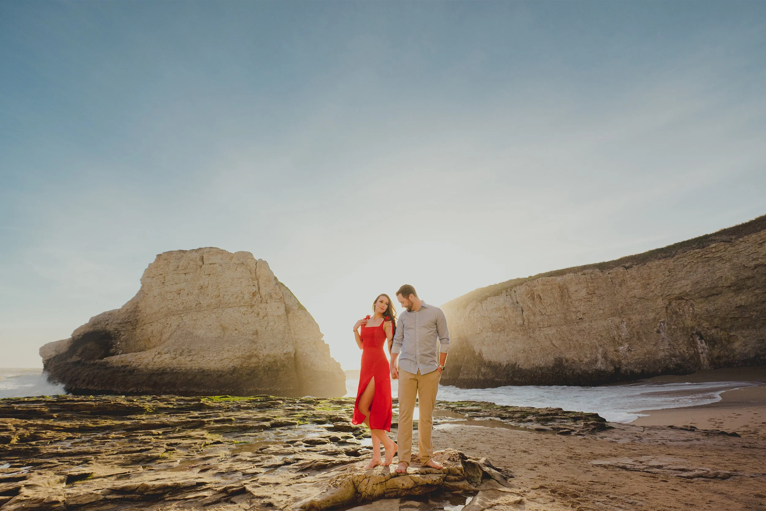Couple with bride-to-be in red dress holding hands near Shark Fin Cove rock formations, award-winning engagement photographer in Orange County