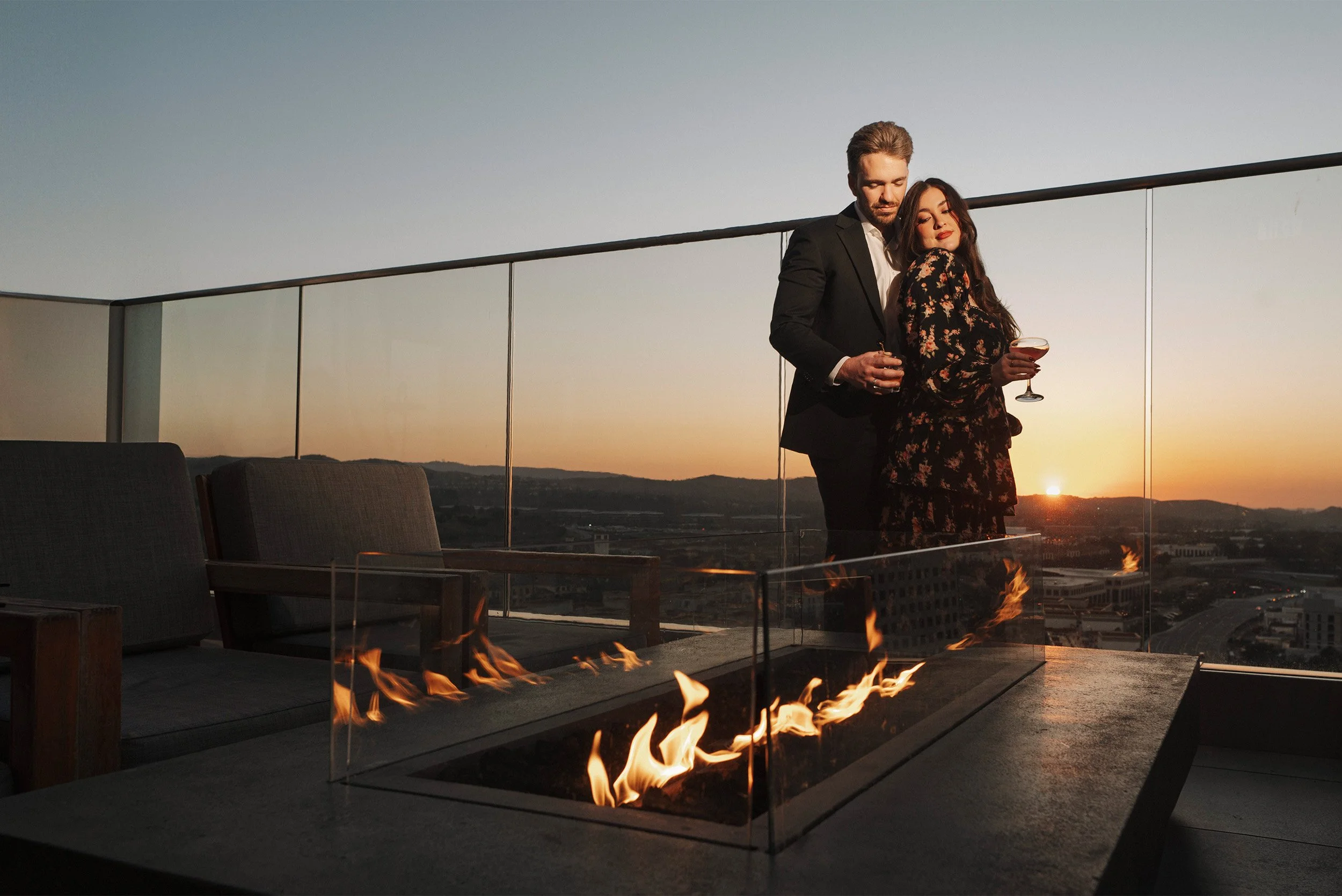 Award-winning engagement photographer in Orange County captures couple sipping cocktails beside a rooftop fire feature at sunset