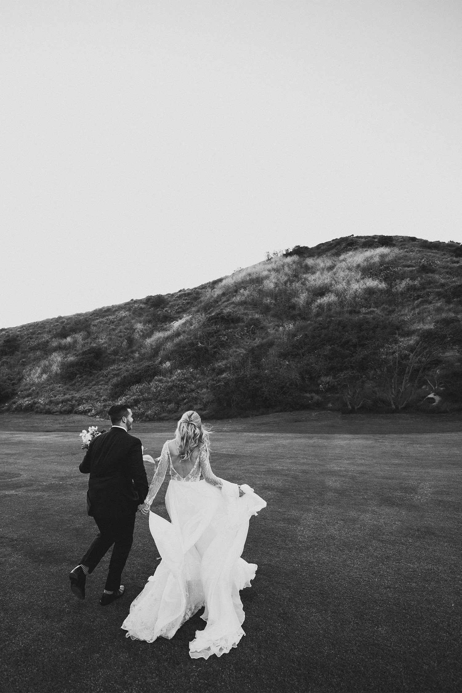 Bride and groom walking hand in hand across open field at The Ranch at Laguna Beach, black and white Orange County wedding photo