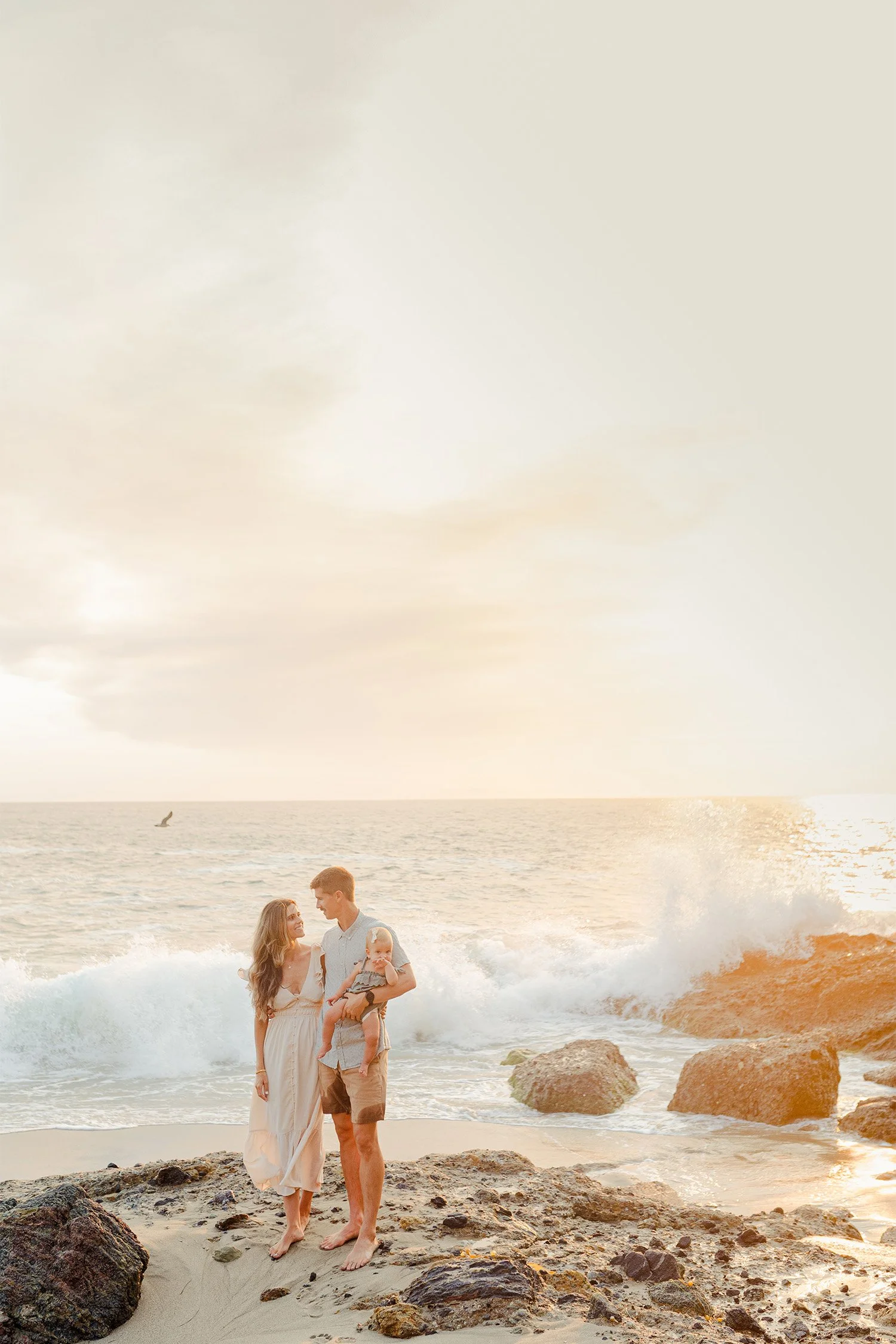 Parents holding their baby on ocean rocks as waves crash at sunset, West coast award-winning family photographer
