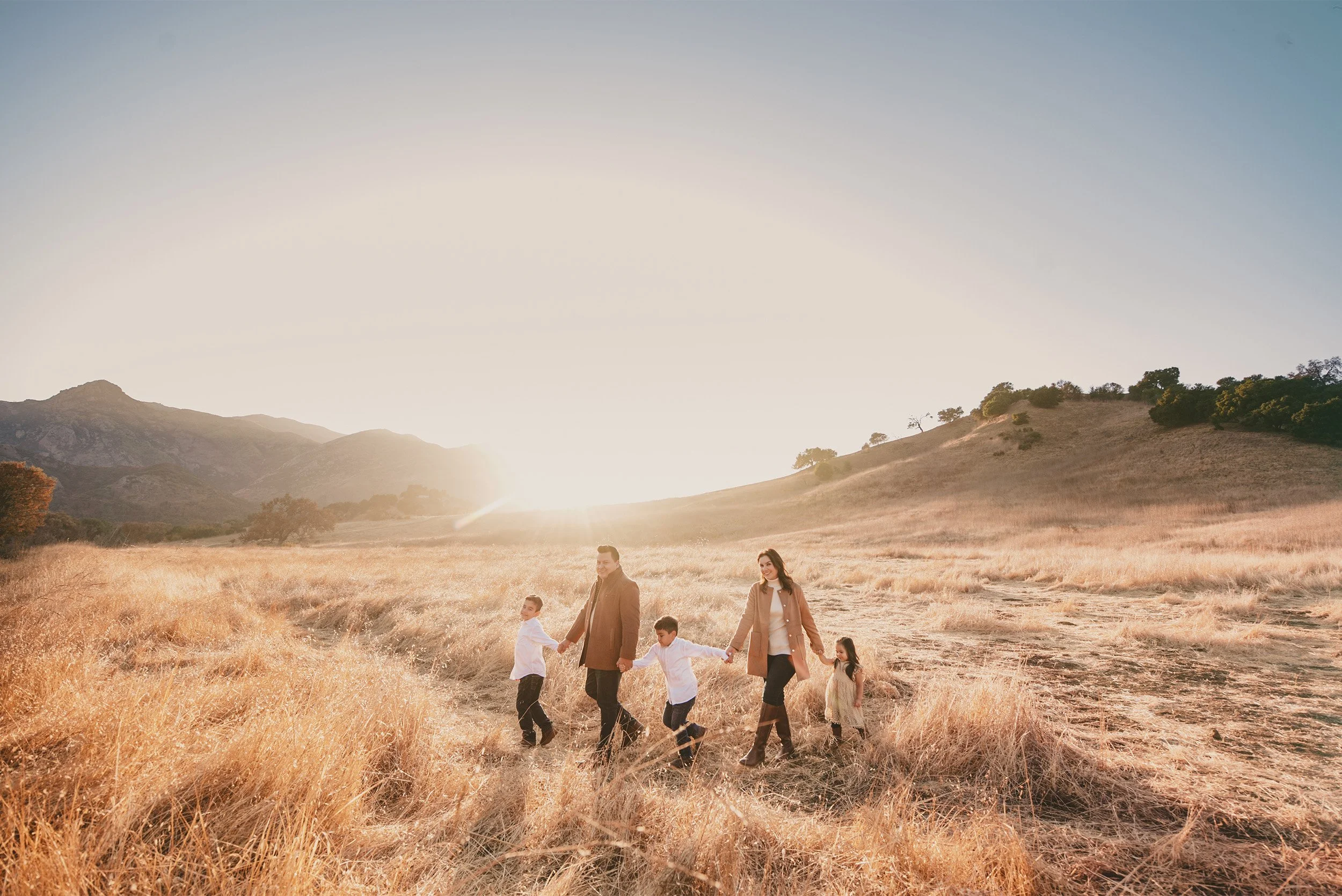 Orange County family photographer photographs family of five walking hand in hand through sunlit grass field at golden hour