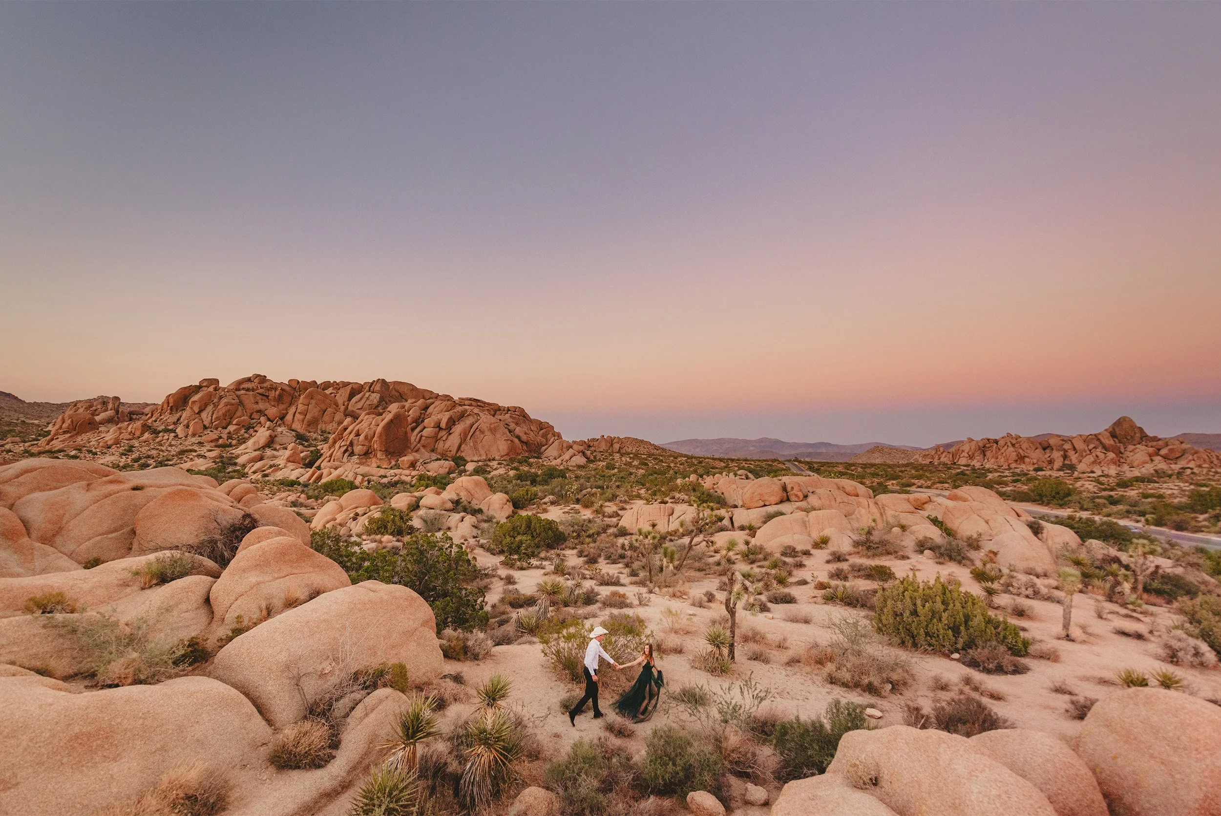 Couple walking hand in hand through Joshua Tree boulders at dusk, desert engagement photos