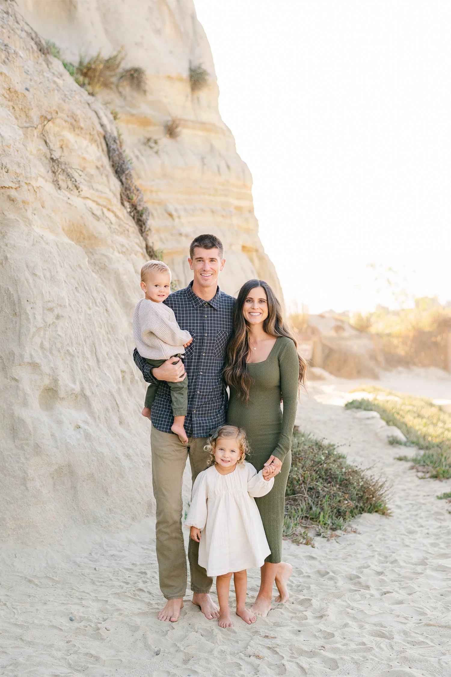 Family of four standing together beneath a sandstone bluff at the beach, Award-winning family photographer in South Orange County