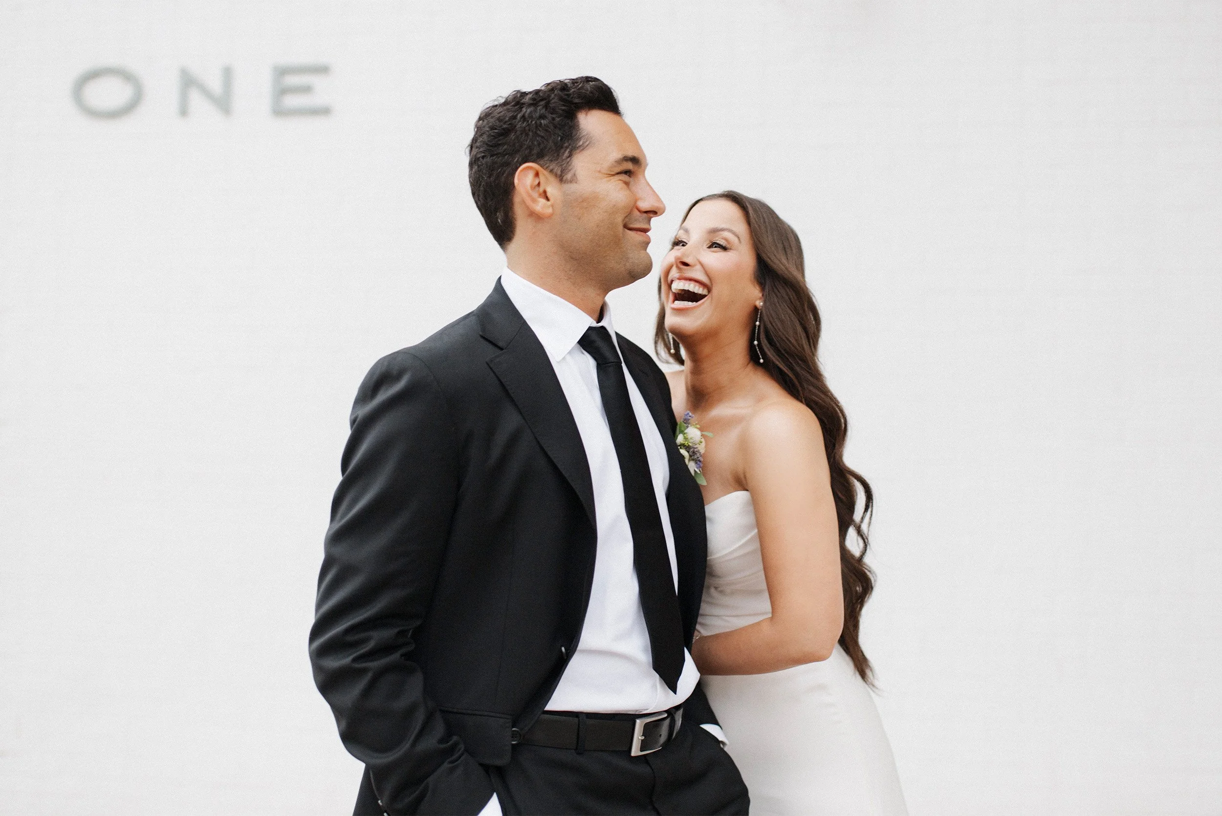 Bride and groom laughing together in front of white wall, candid Orange County wedding portrait