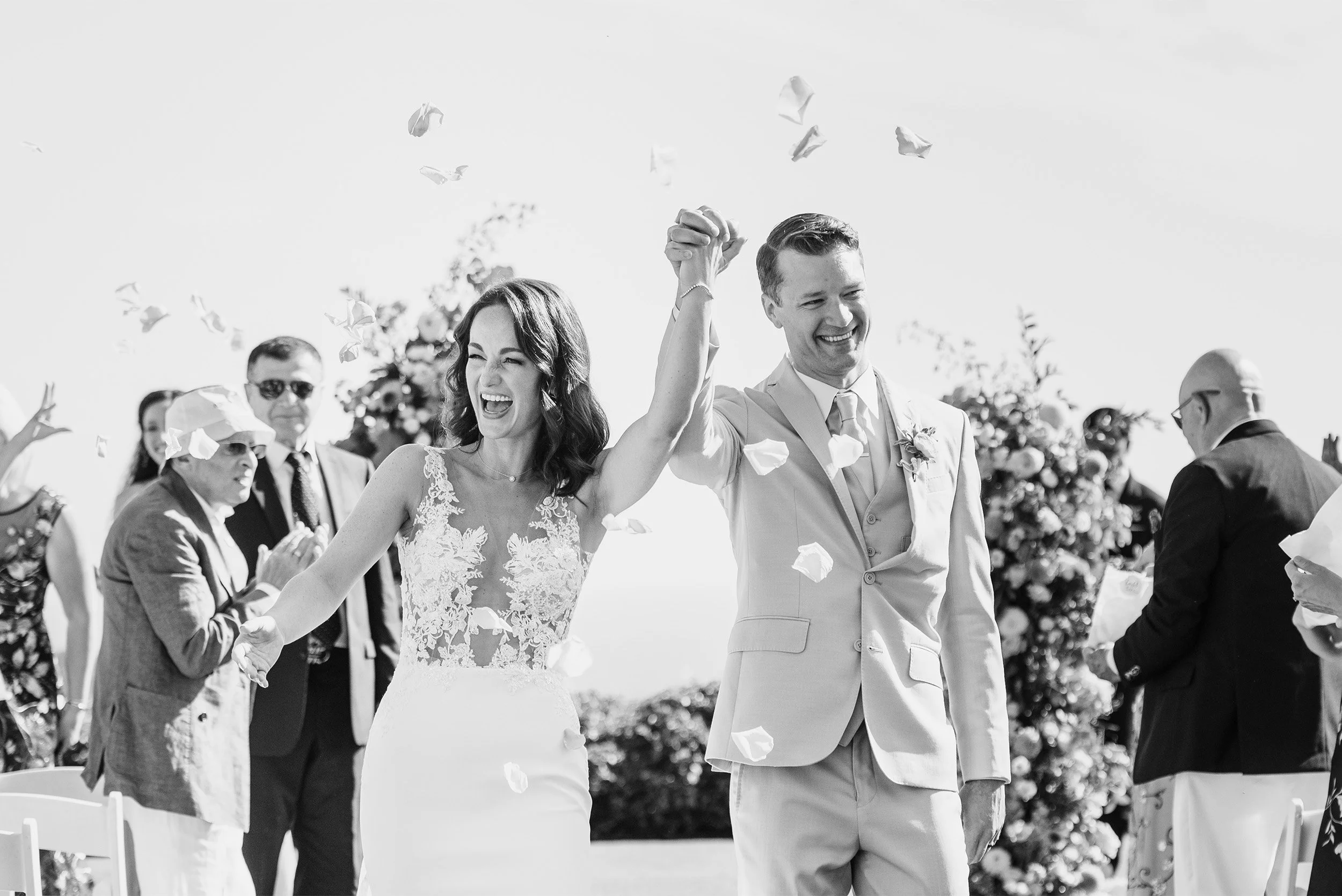 Highly recommended wedding photographer in Santa Barbara  captures bride and groom celebrating with raised hands during flower petal recessional, black and white