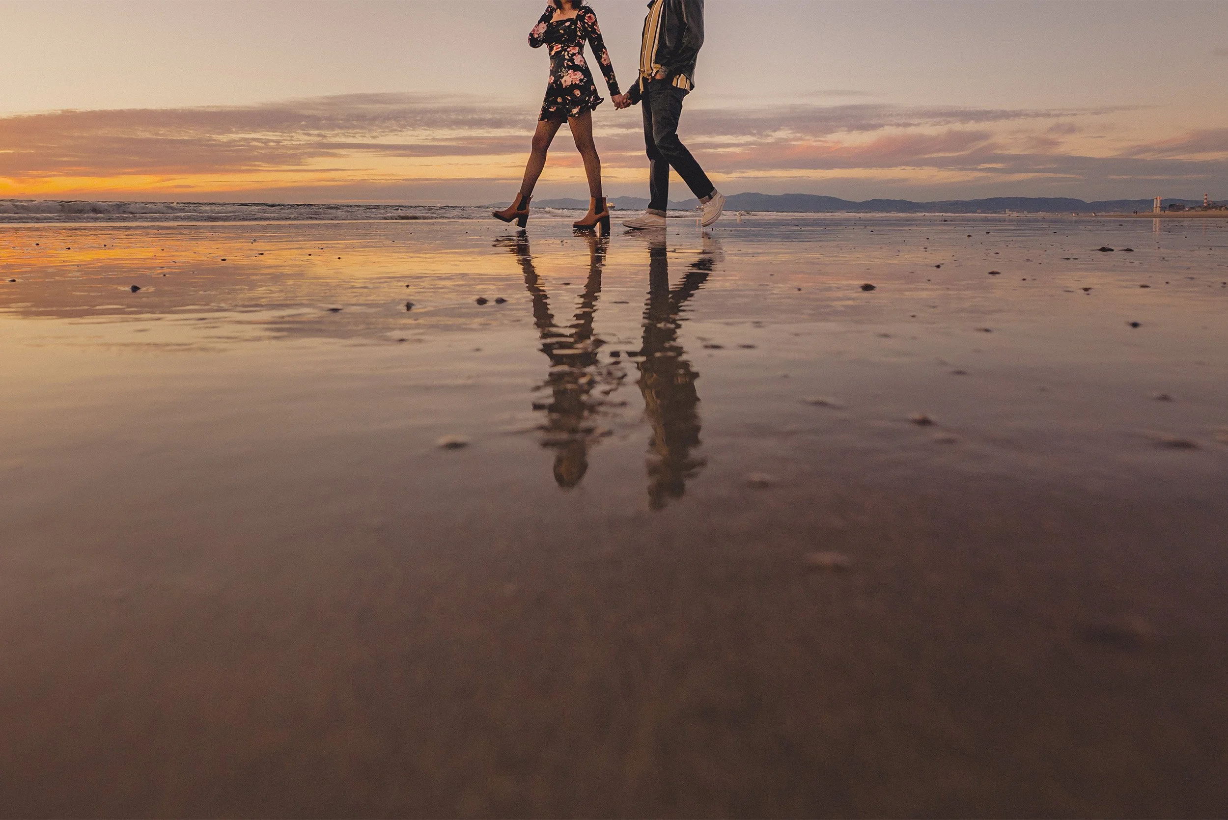 Award-winning engagement photographer at Manhattan Beach captures couple walking hand in hand along the reflective shoreline at sunset