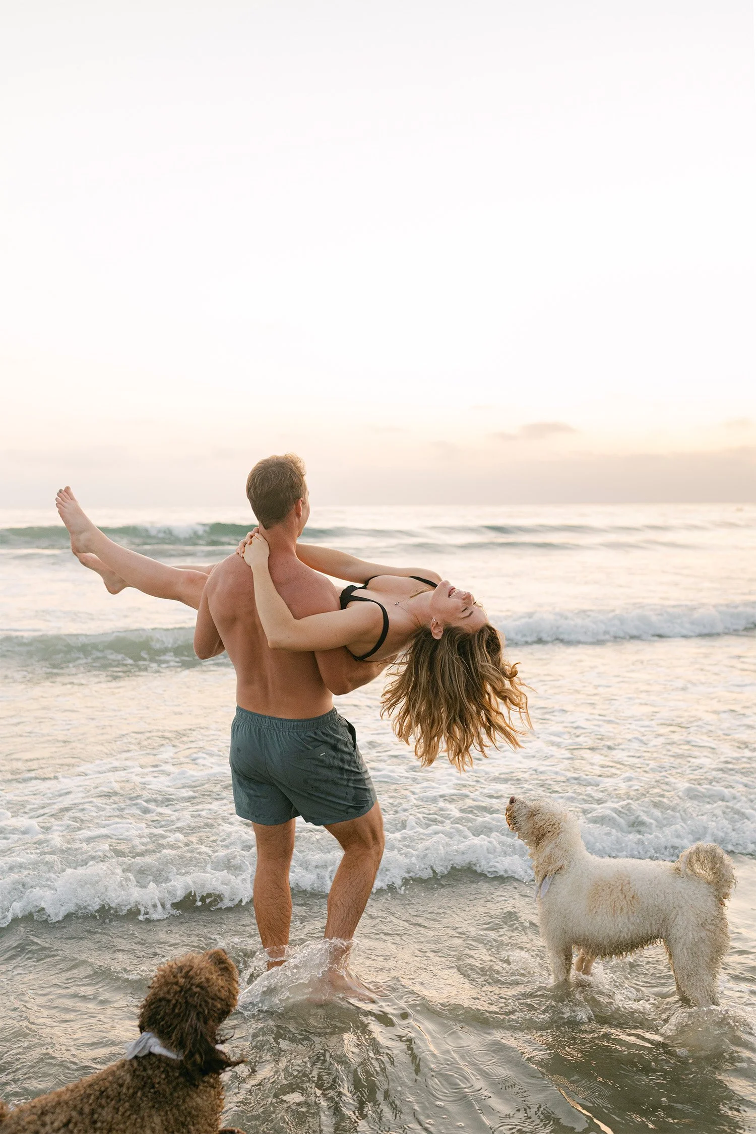 Man lifting pregnant partner playfully at beach with dogs, Orange County maternity photographer
