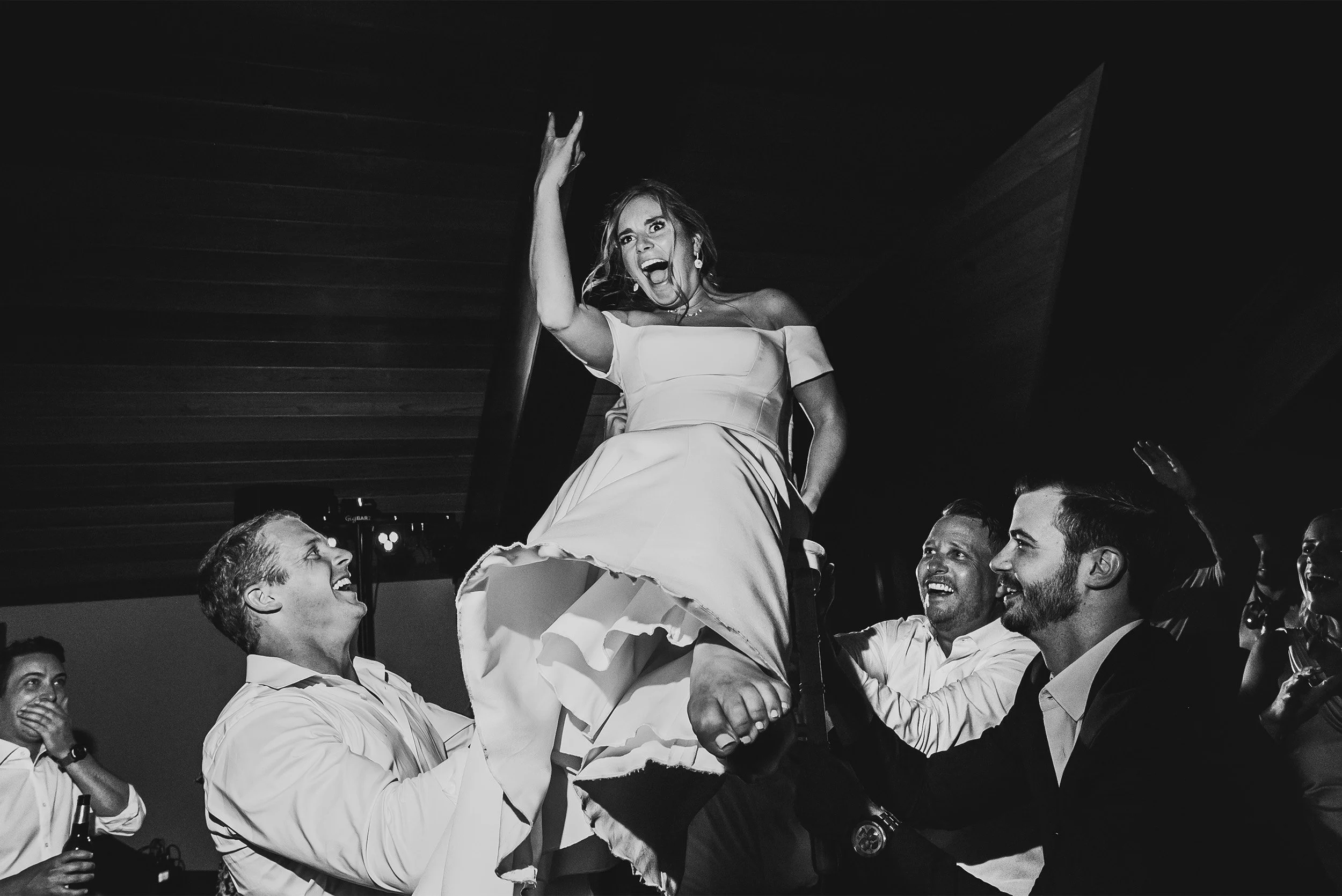 Bride lifted on shoulders celebrating during wedding reception dance floor, black and white photo