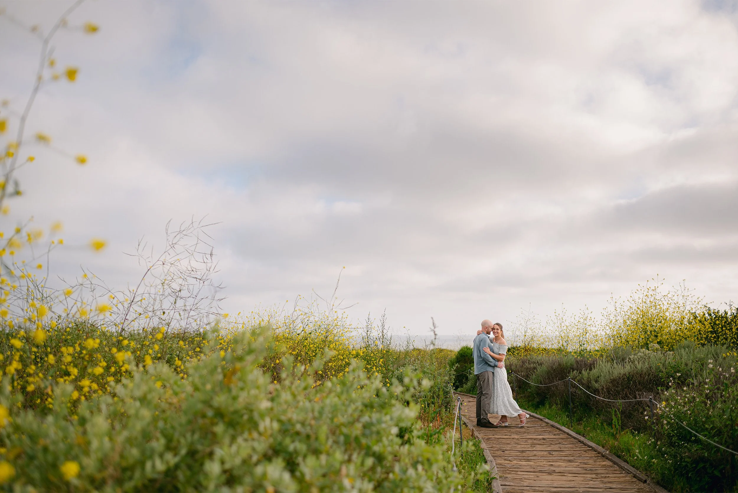 Highly recommended engagement photographer in Orange County captures couple embracing on the Crystal Cove boardwalk lined with wildflowers