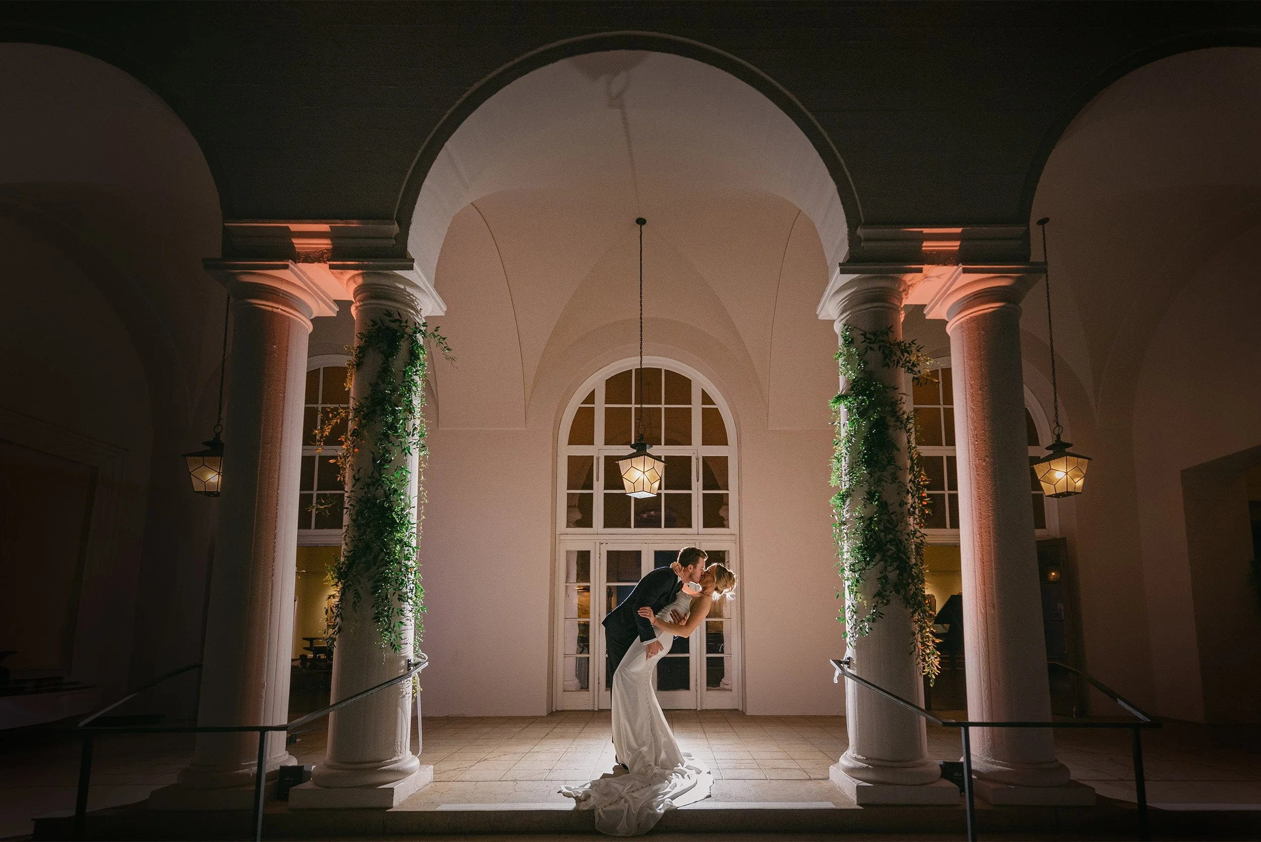 Highly recommended wedding photographer in Orange County captures romantic nighttime kiss beneath illuminated stone archway with hanging lanterns at The Ebell of Los Angeles