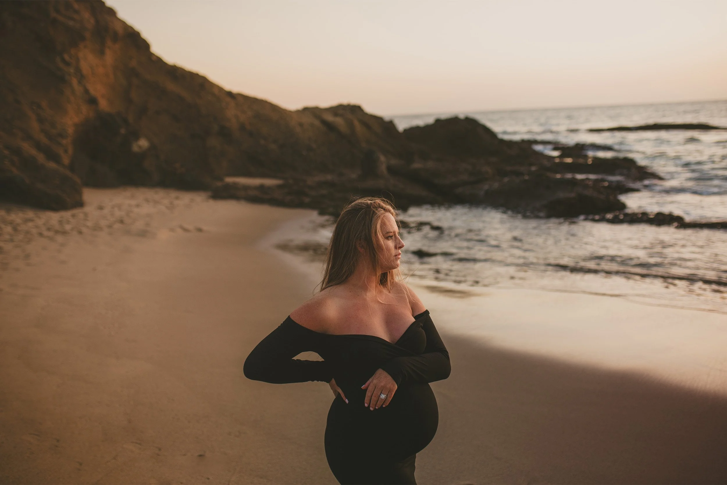 Orange County maternity photographer captures pregnant woman in black gown on rocky beach at sunset