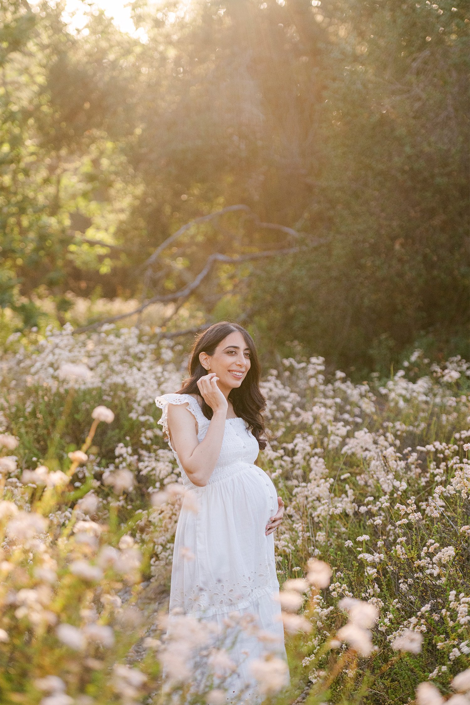 Pregnant woman smiling in white dress in golden wildflower field, highly recommended maternity photographer in Orange County