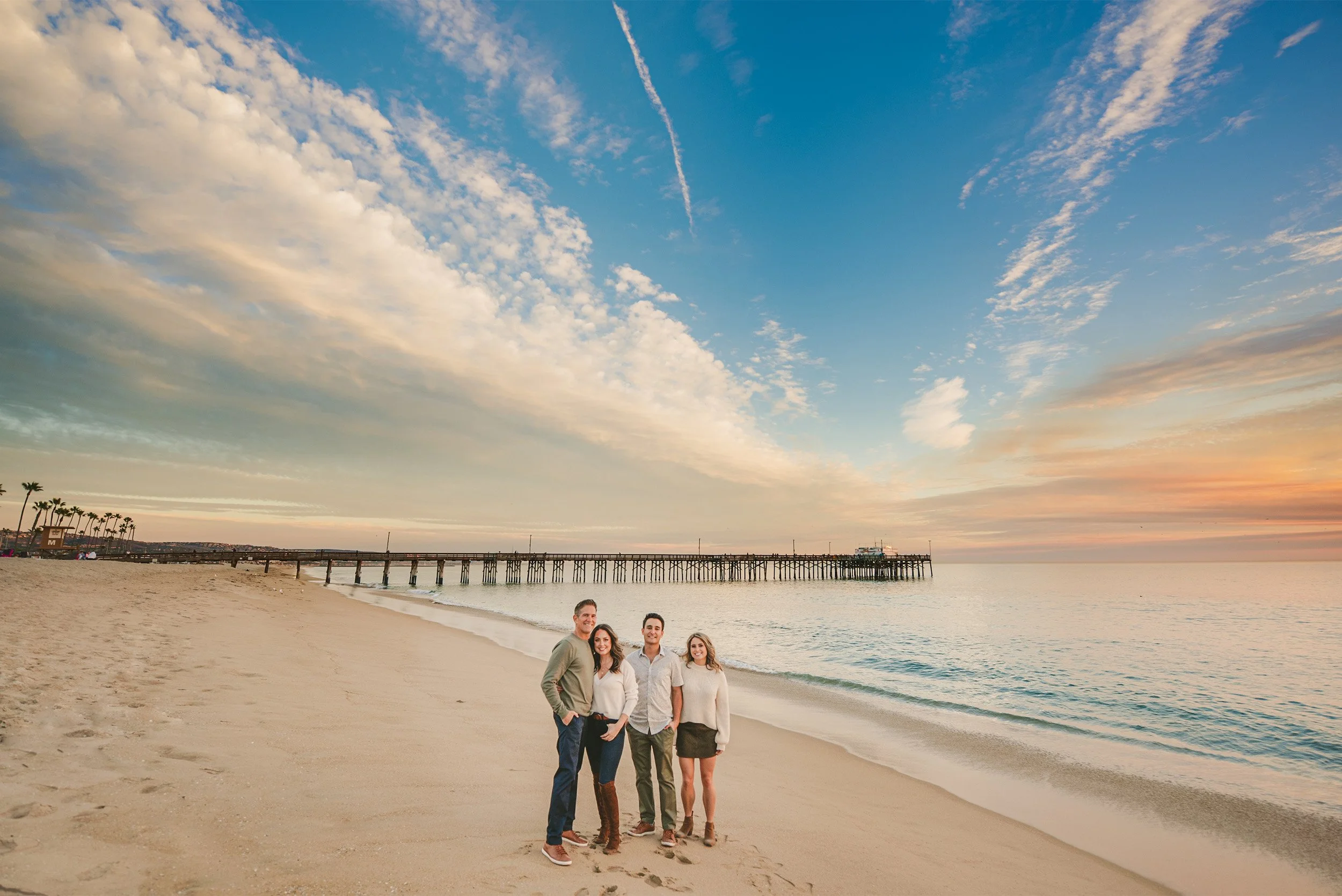 Orange County family photographer captures family portrait on beach at sunset near pier with dramatic sky