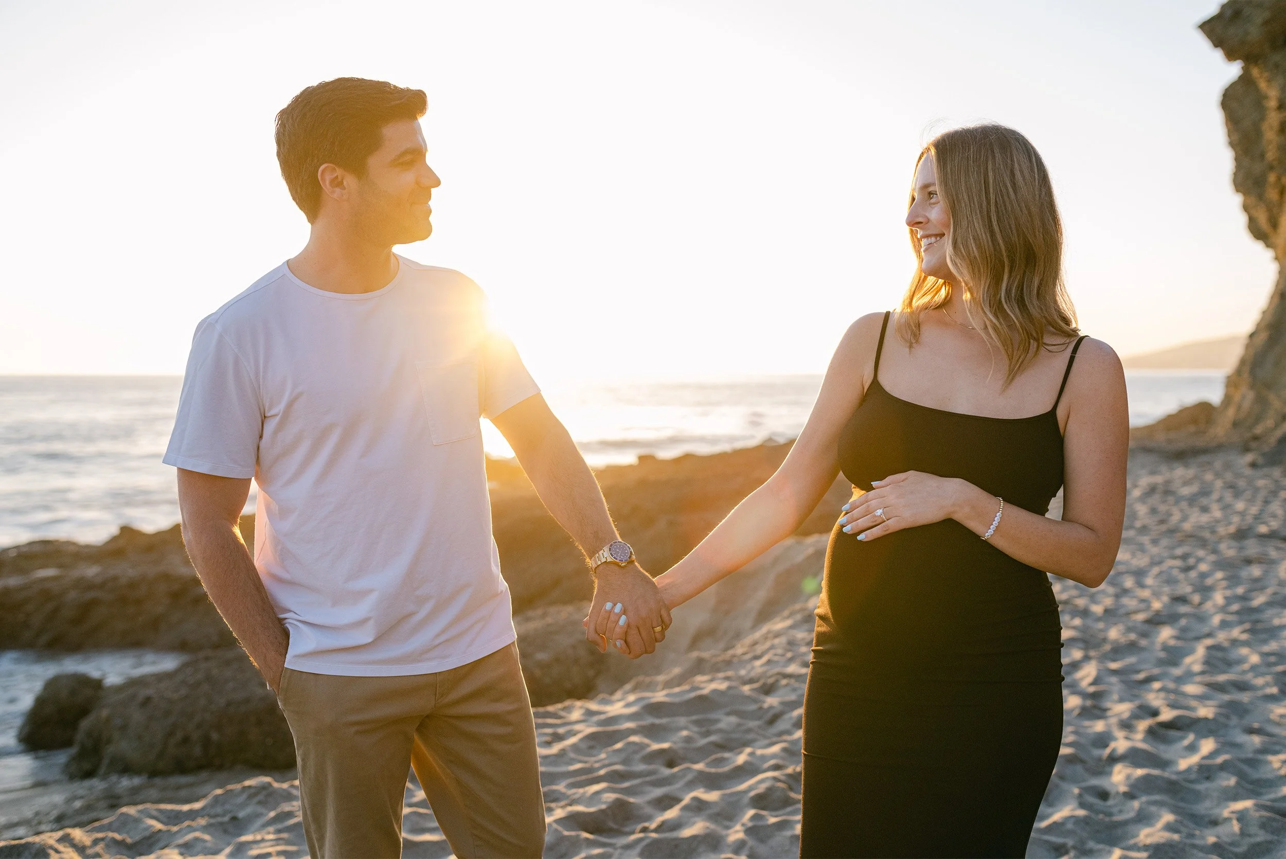 Pregnant woman in black dress holding hands with partner on beach, Orange County maternity photographer