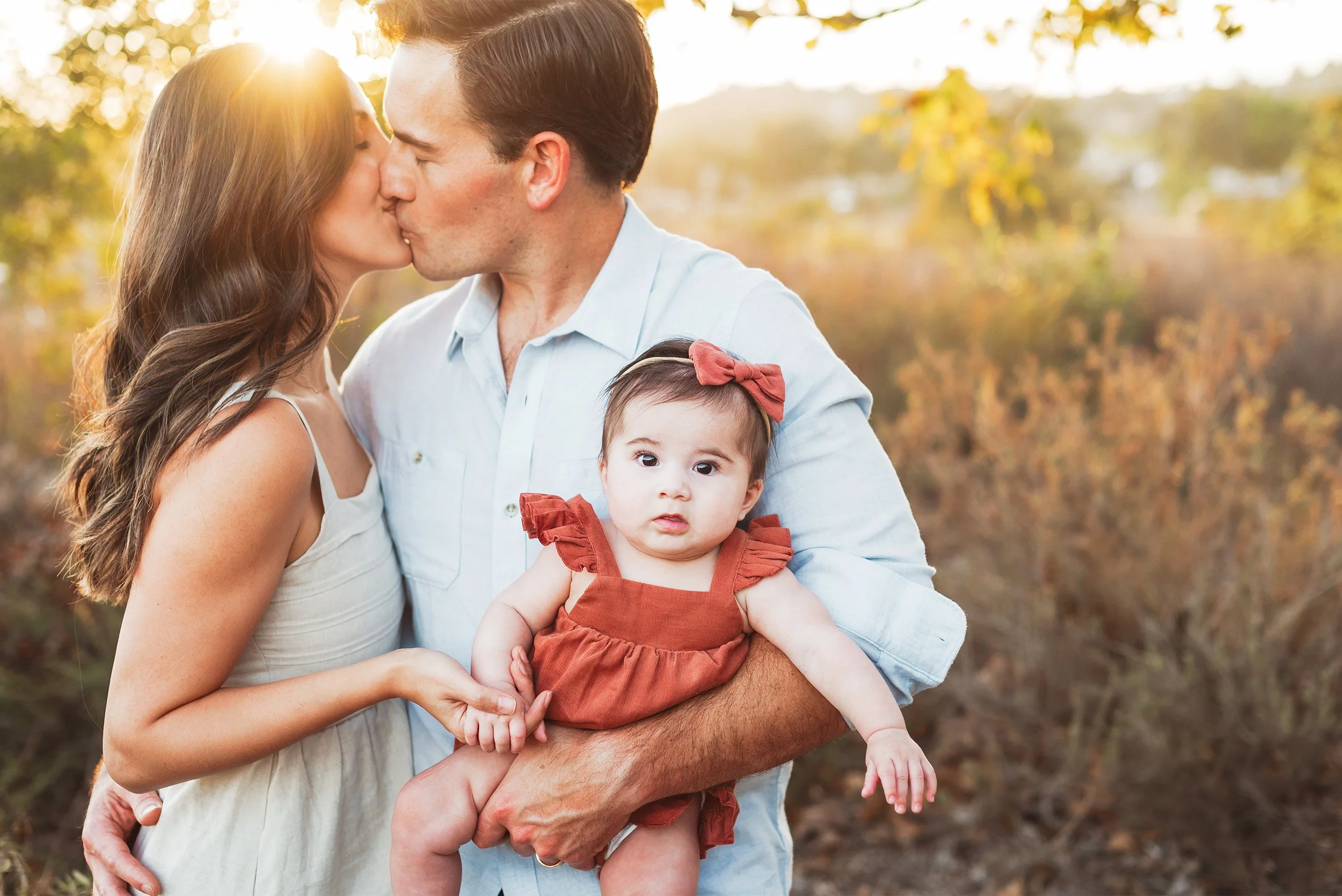 Orange County family photographer captures parents sharing a kiss while dad holds baby girl in rust-colored romper at golden hour