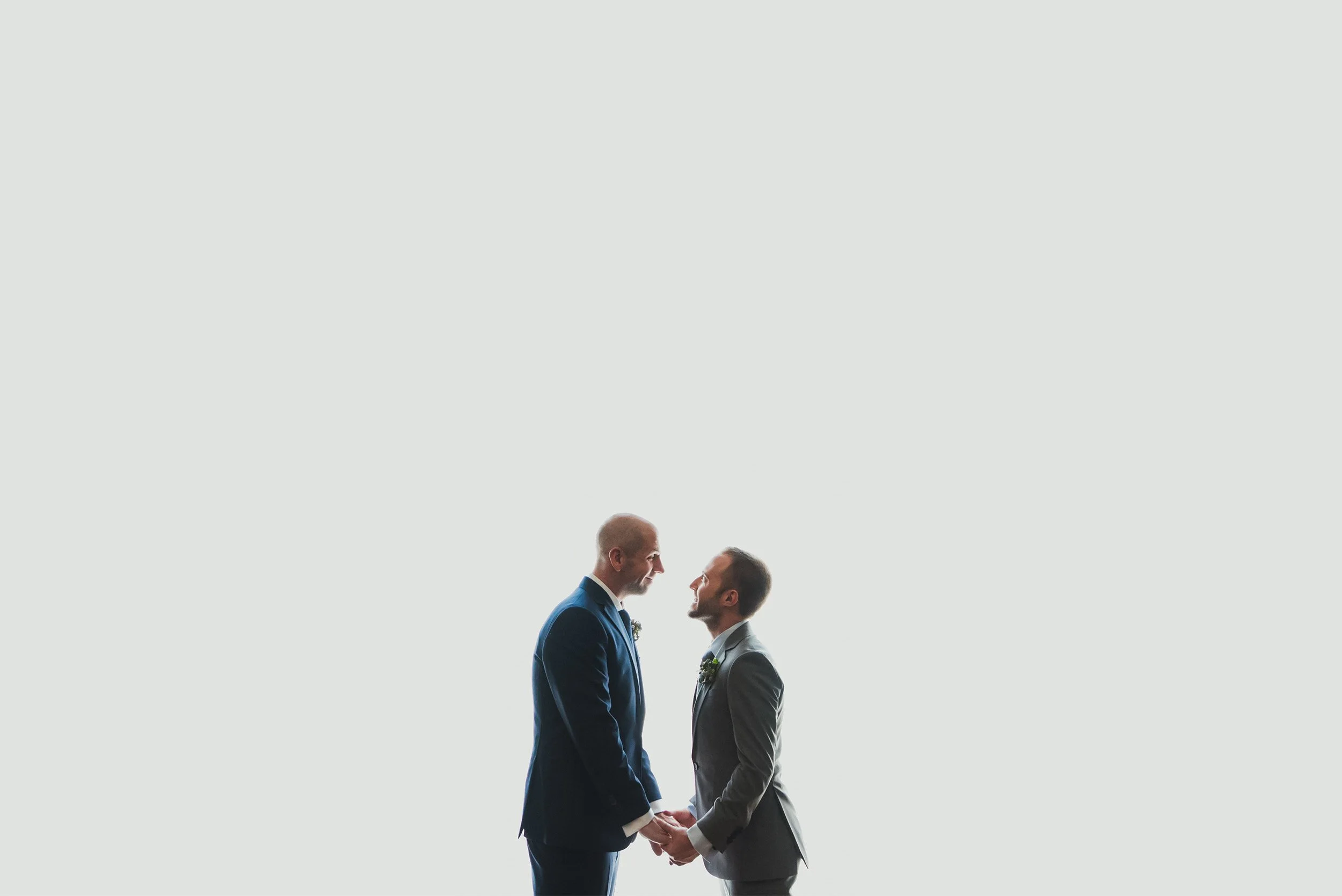 Two grooms holding hands and smiling at each other, minimalist LGBTQ+ wedding portrait