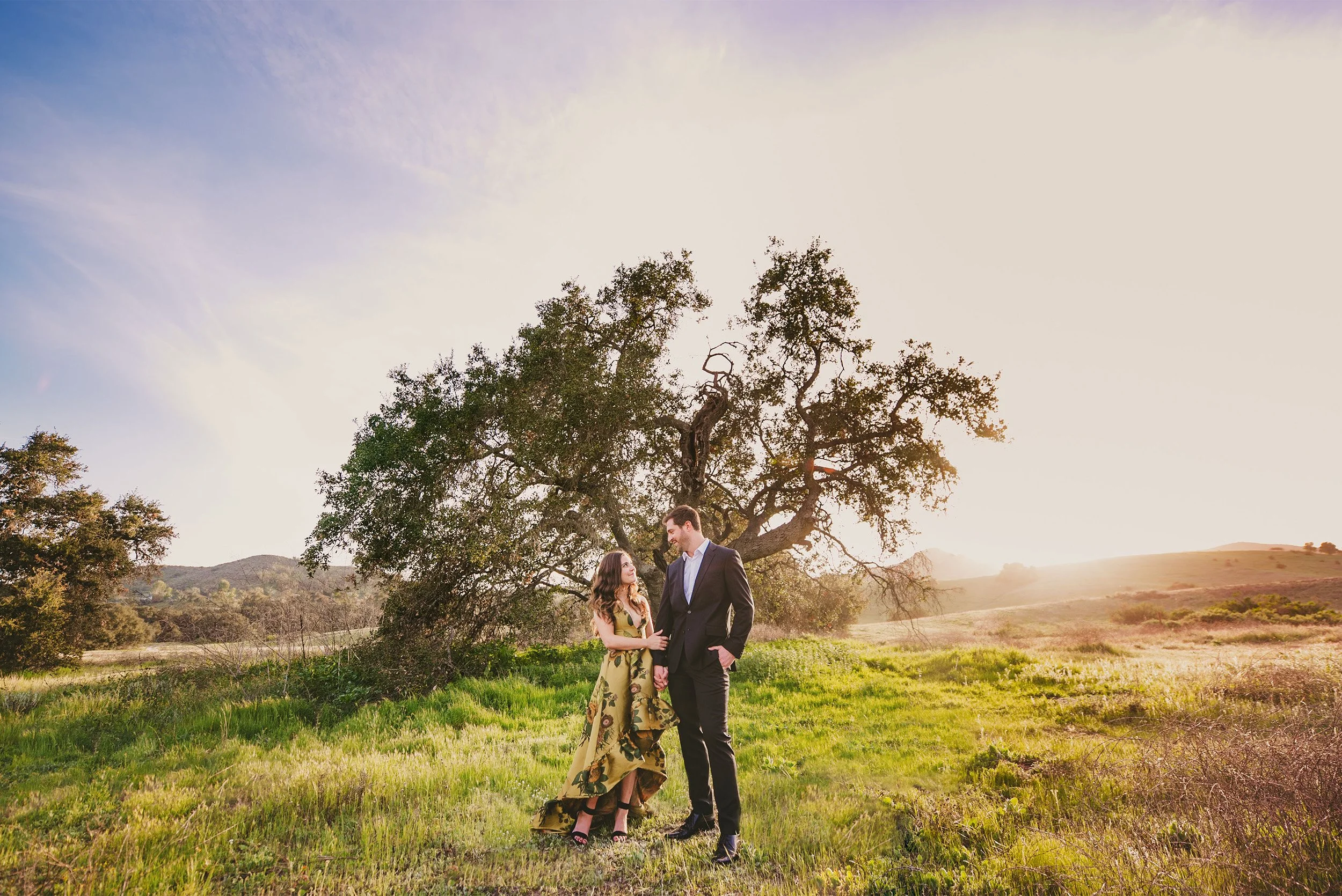 Bride-to-be in a floral dress and groom-to-be under an oak tree at sunset, Southern California engagement portrait