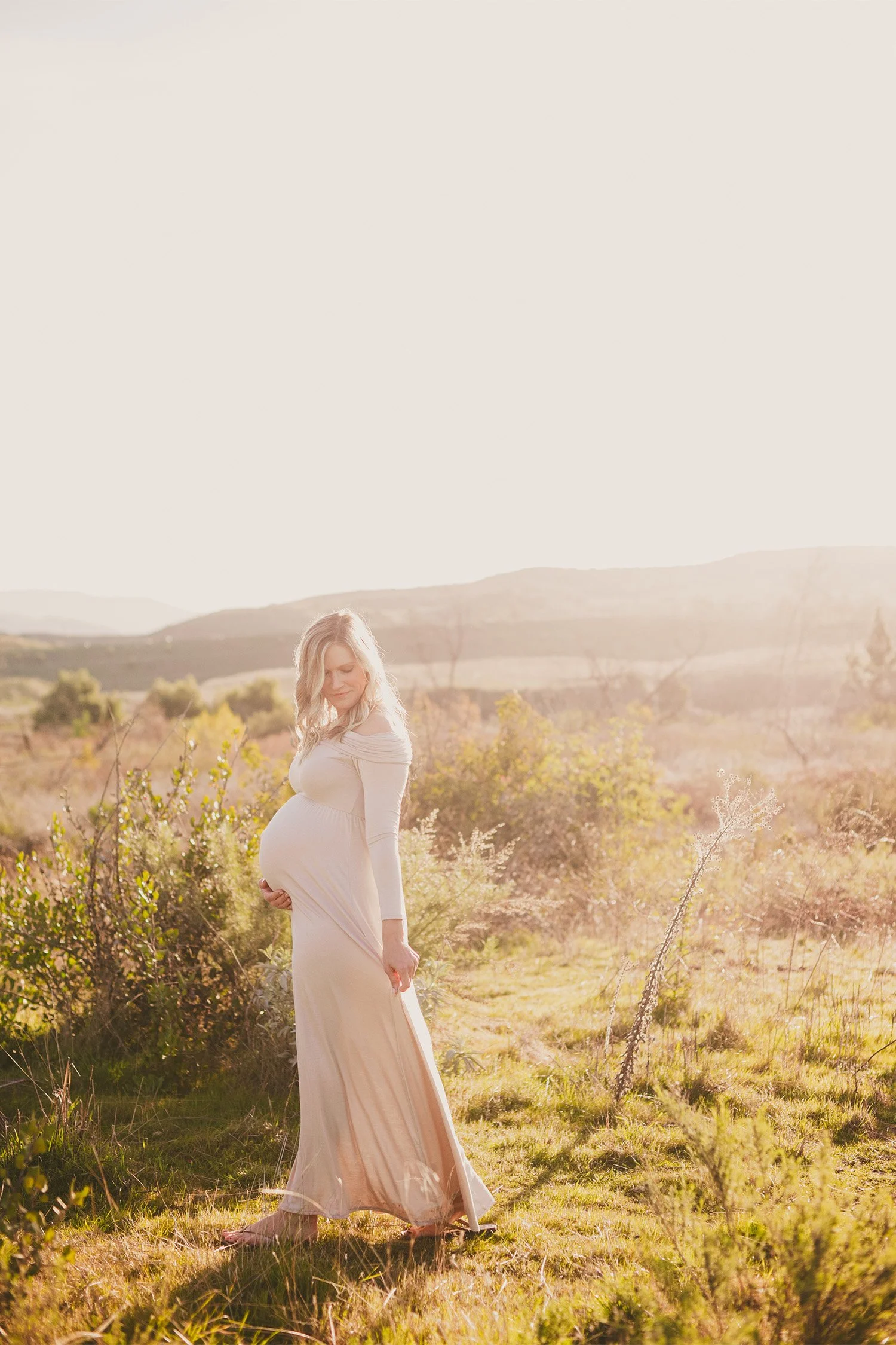 Orange County maternity photographer photographs pregnant woman in flowing cream gown walking through golden field at sunset