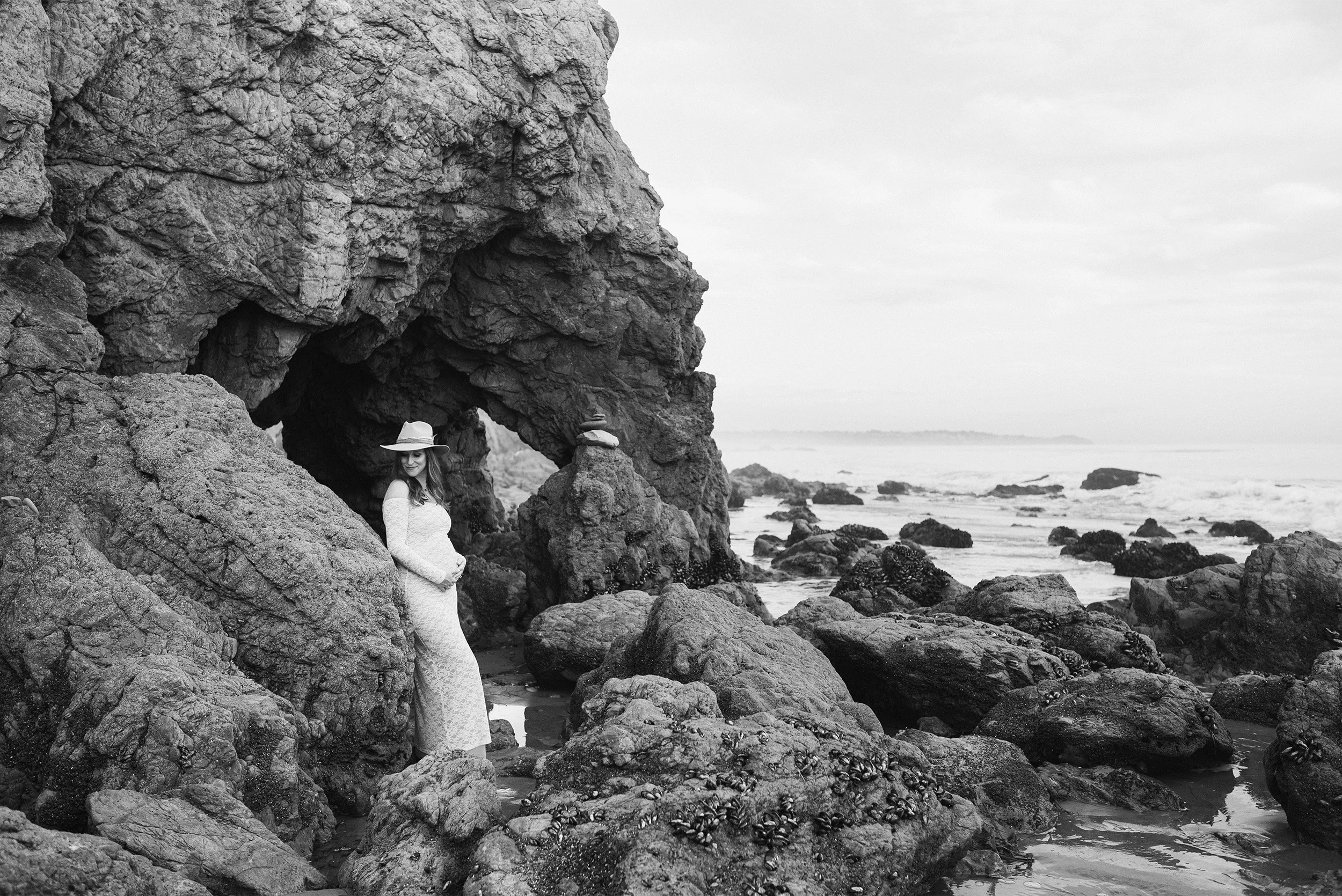 Black and white photo of pregnant woman standing in rocky sea cave, highly recommended maternity photographer Orange County