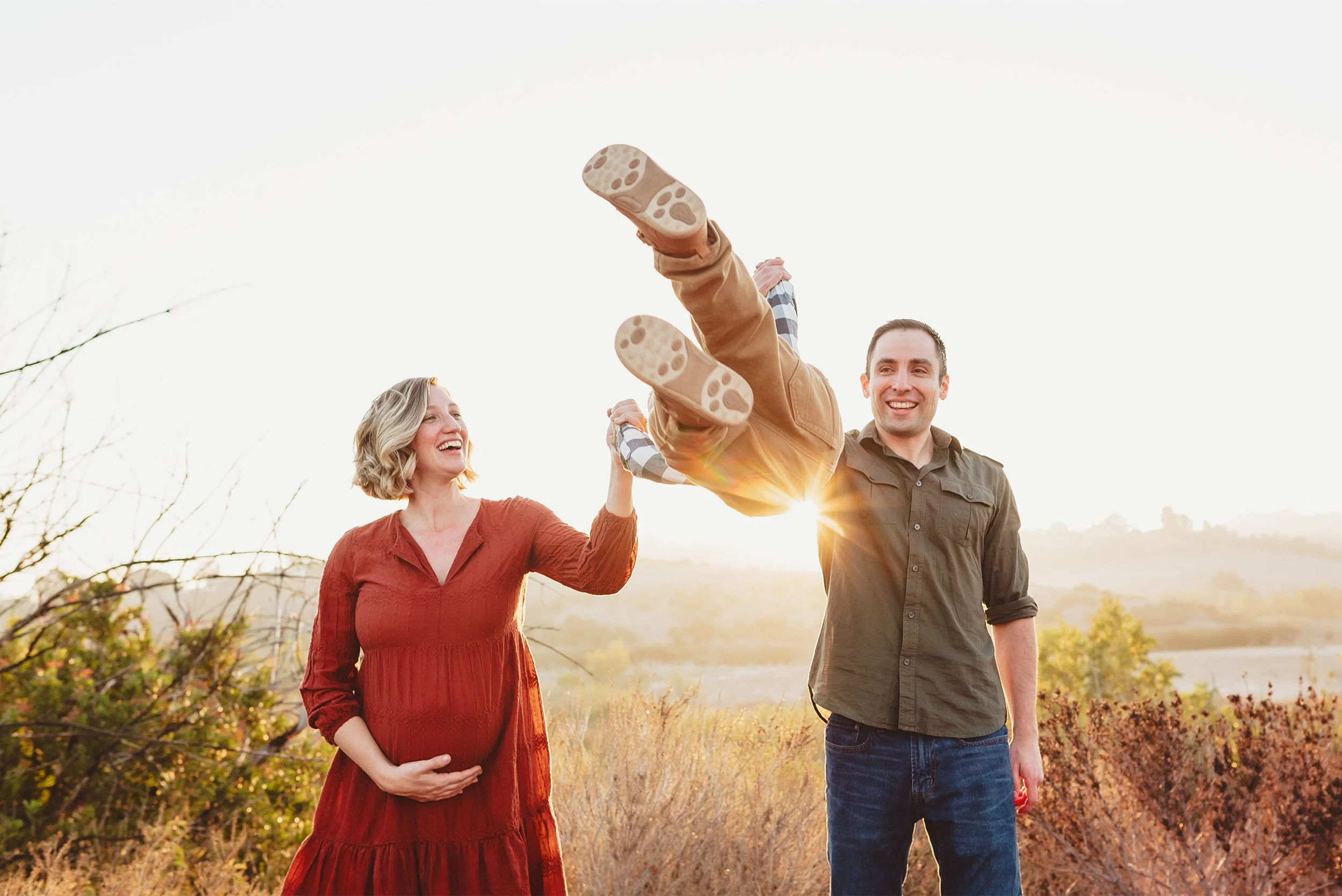 Orange County maternity photographer and family photographer captures pregnant mother and father playfully lifting toddler in golden field