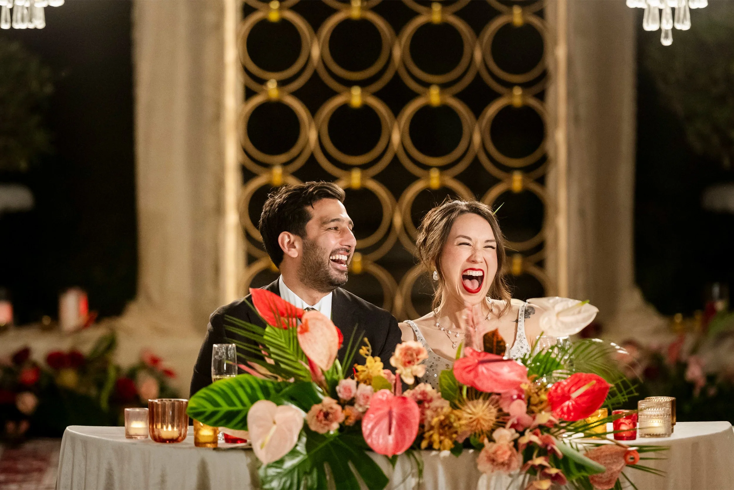 Award-winning wedding photographer in Orange County captures bride and groom laughing together at reception sweetheart table with tropical florals, South Asian Wedding