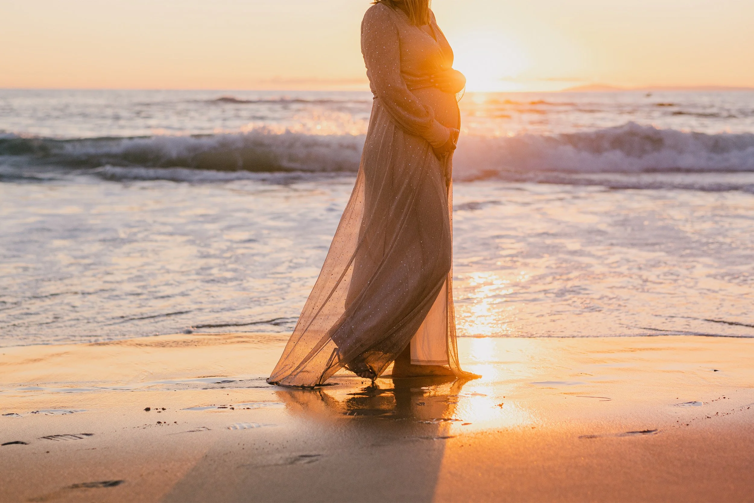 Silhouette of pregnant woman in flowing dress standing at shoreline, highly recommended maternity photographer Orange County