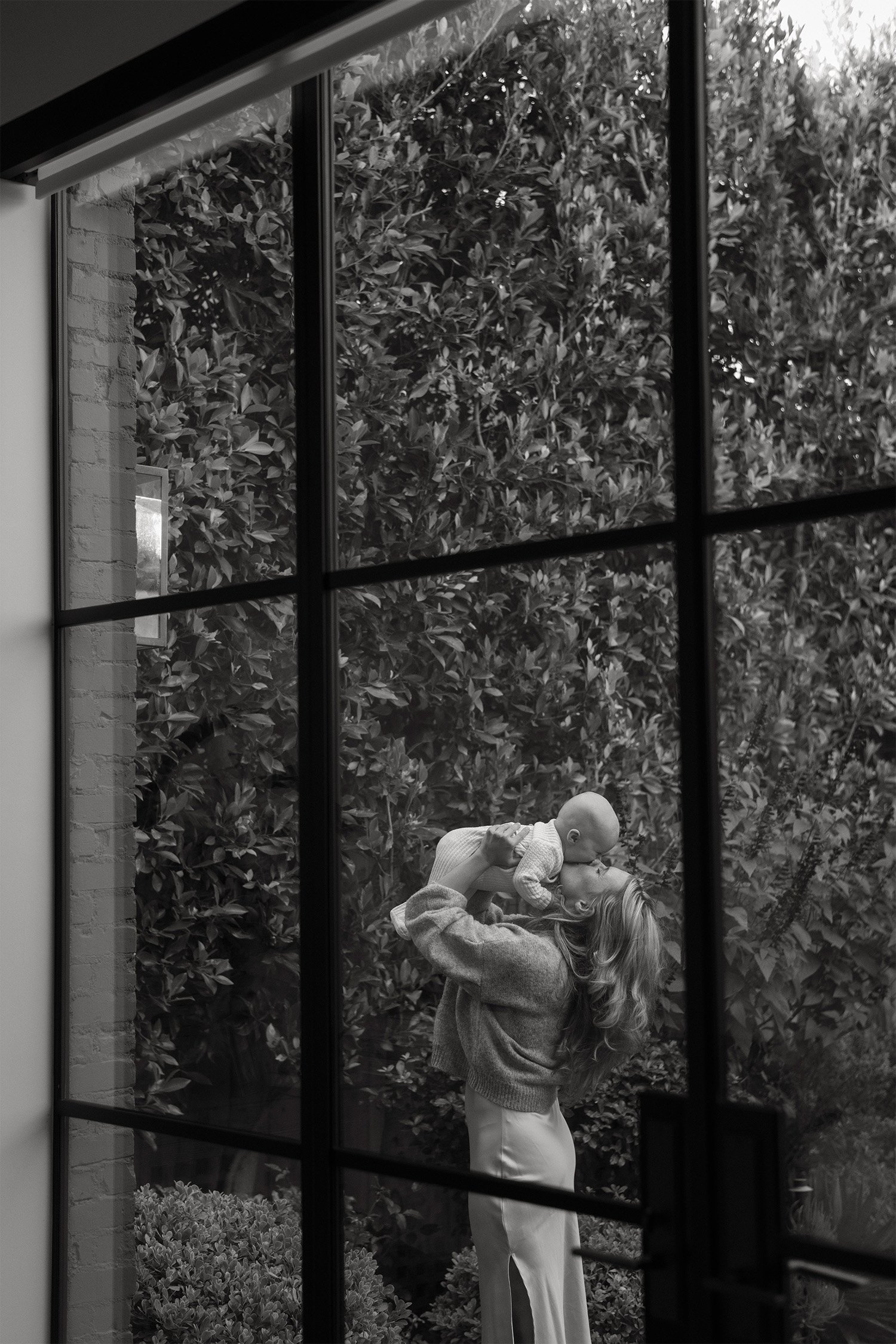 Orange County family photographer captures mother lifting baby up through a garden window reflection, in home family photos