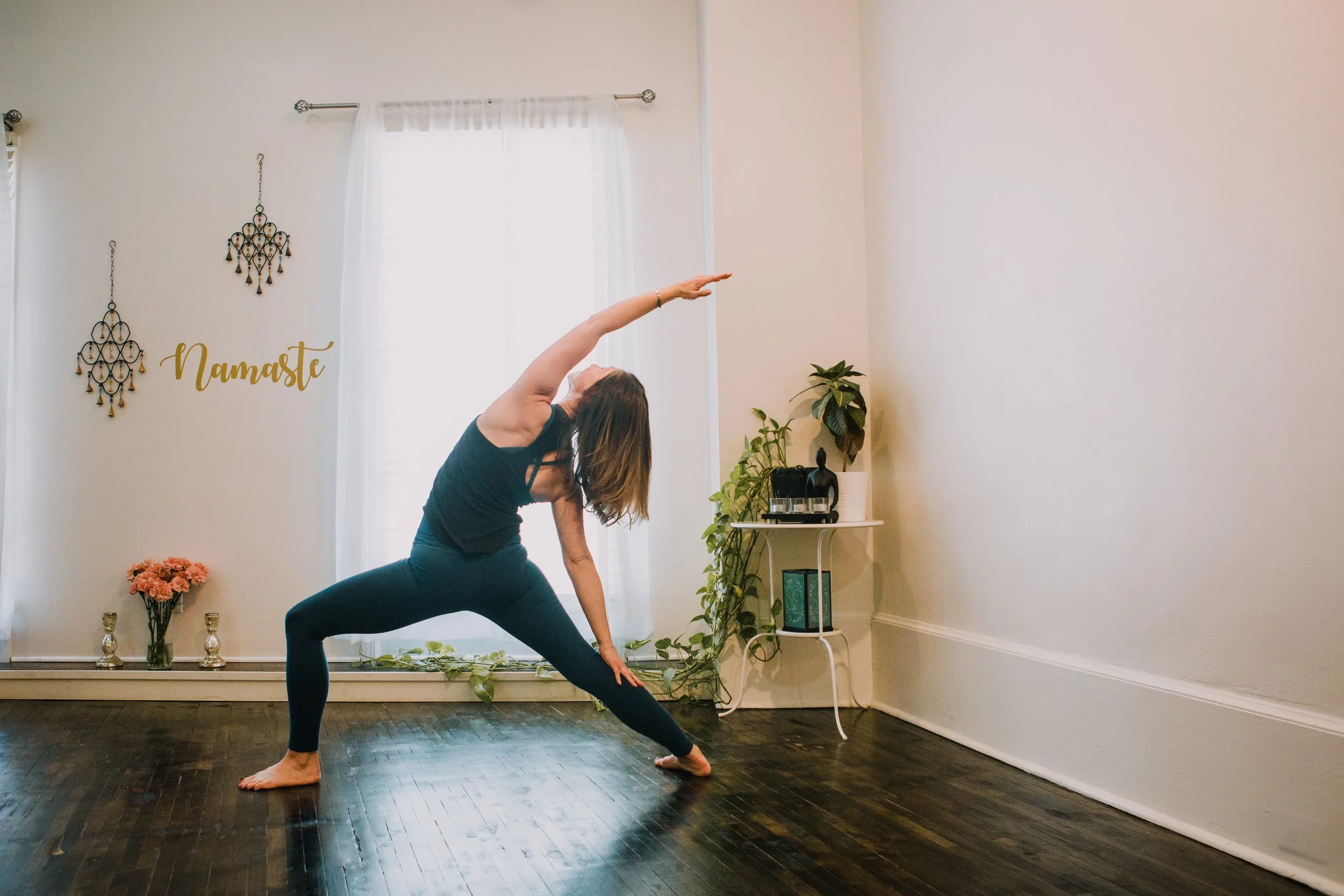 Person practicing yoga in a serene room with decorative elements, including wall hangings, flowers, and plants, with natural light filtering through windows.