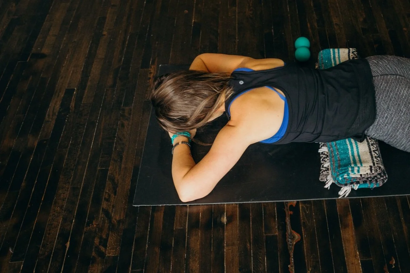 Person lying face down on a yoga mat with a blanket and massage balls on a hardwood floor.