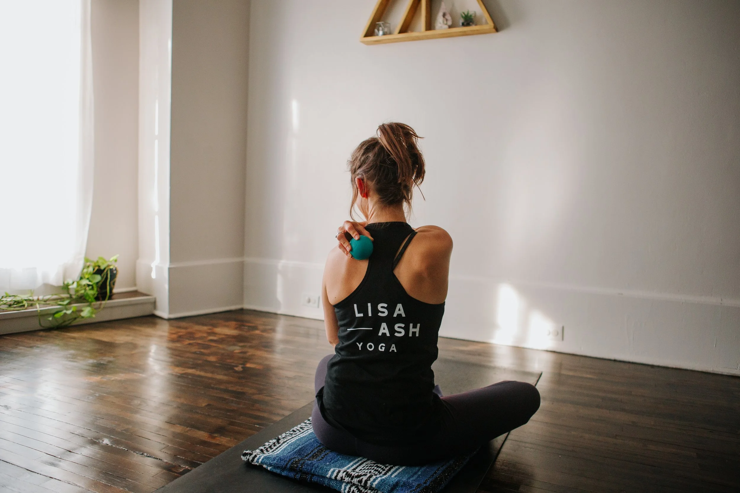 Person sitting on a yoga mat with text 'Lisa Ash Yoga' on their tank top, holding a massage ball to their shoulder, in a bright room with wooden floors.