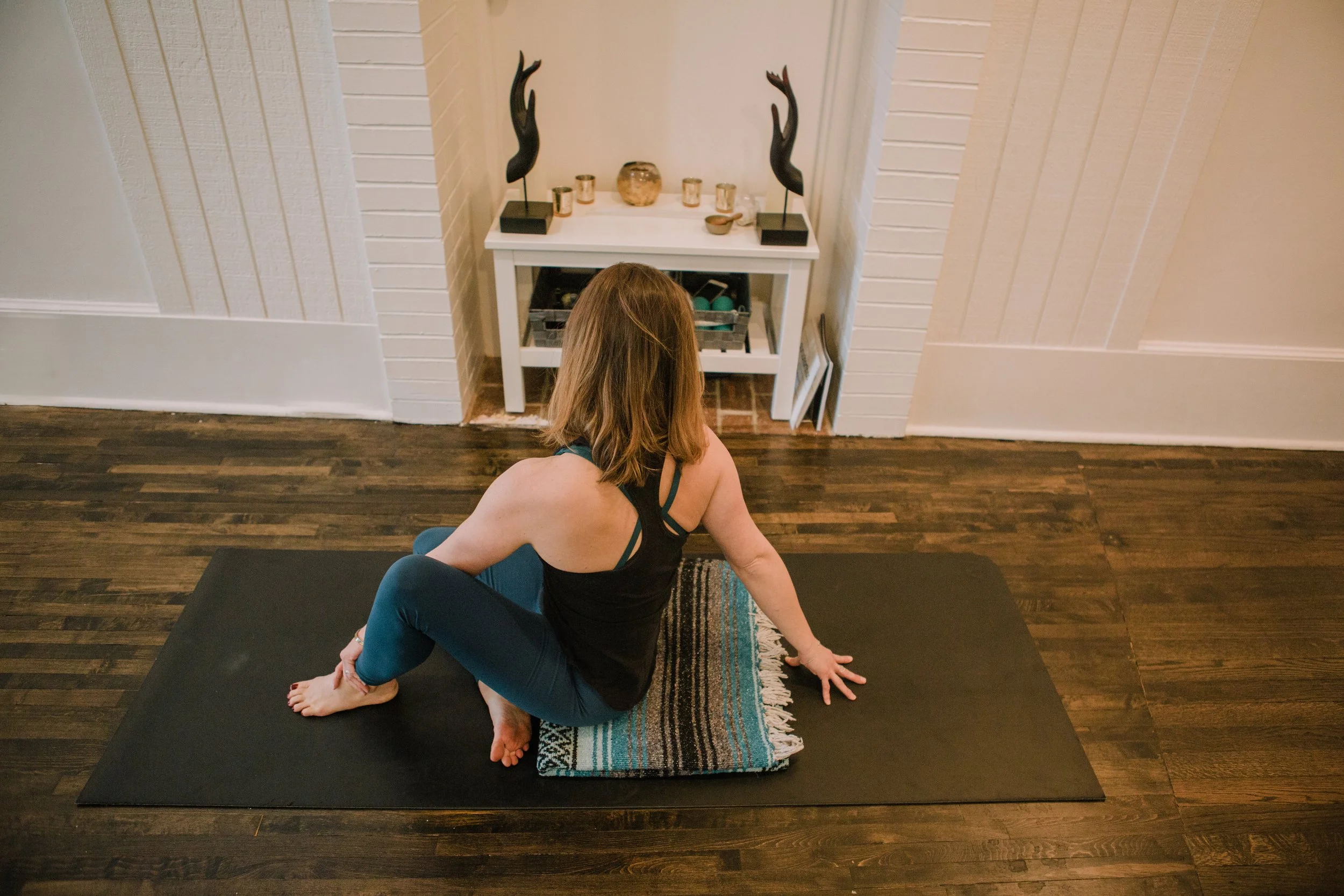 Woman practicing yoga at home on a black mat, sitting cross-legged with one hand on the floor, wearing a black tank top and blue leggings, with a decorative table in the background.