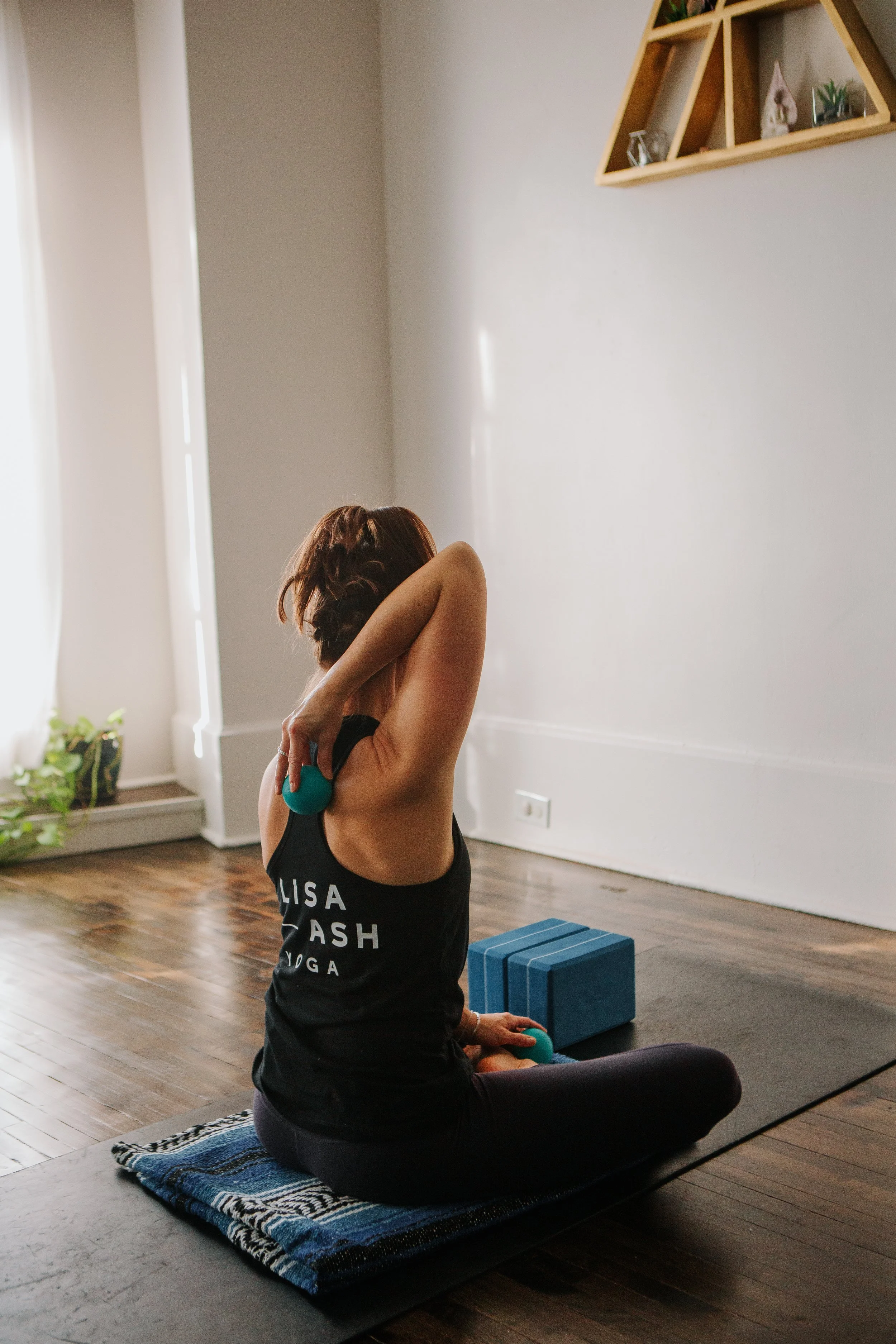 Woman sitting on a yoga mat using massage balls, with yoga blocks nearby, in a serene room.