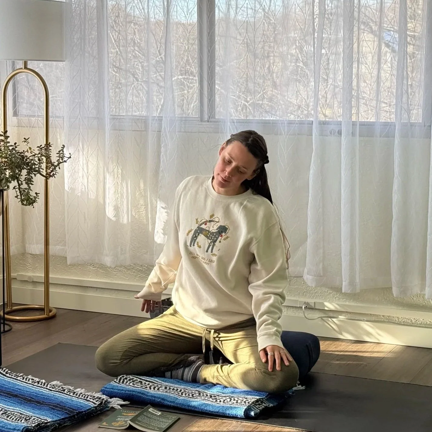 A woman practicing yoga on a blue mat on wooden floor, in front of a wooden bench with a laptop showing a yoga video, a yellow toy bulldozer, and a green toy car nearby.