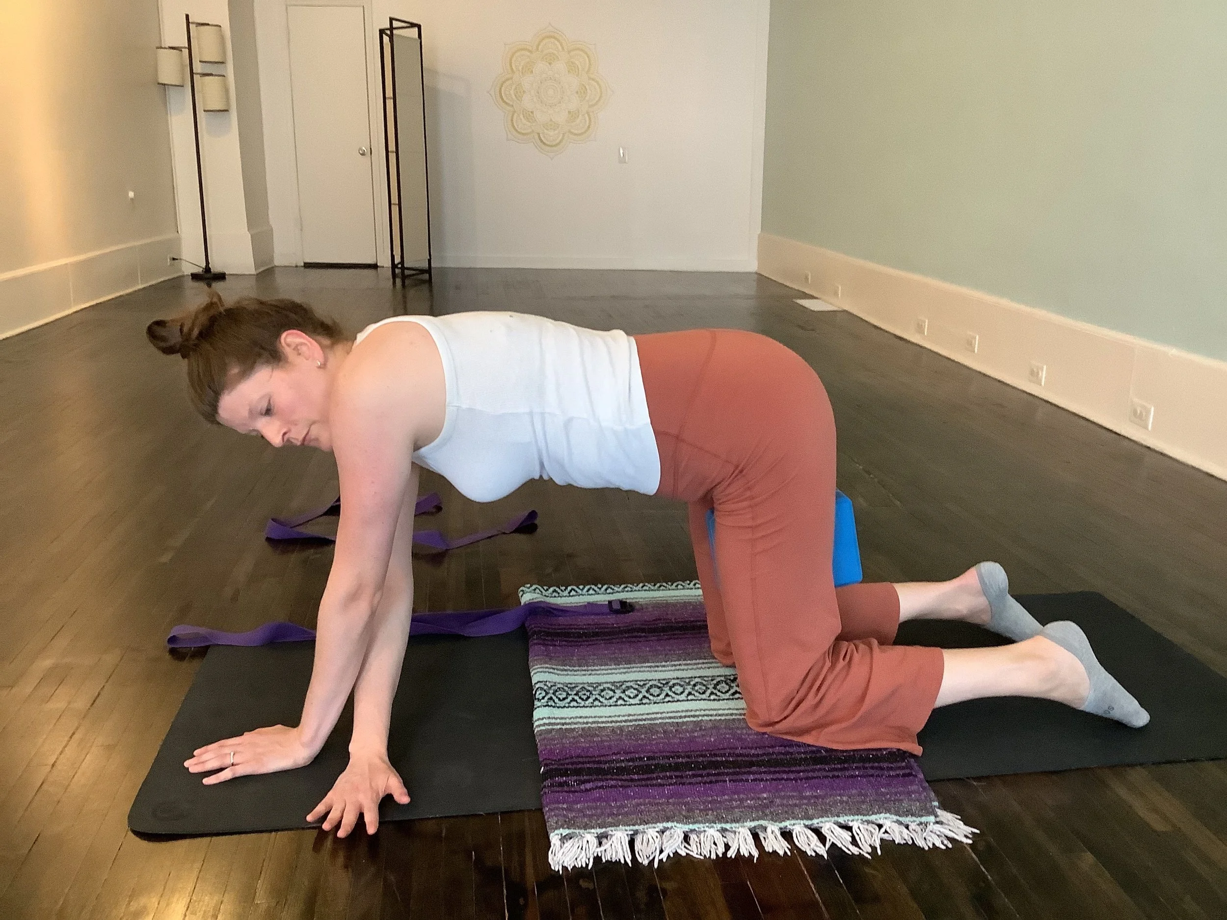 Woman practicing yoga on a mat indoors, wearing a white tank top and brown pants, with ankles crossed and resting on a purple and black patterned blanket.