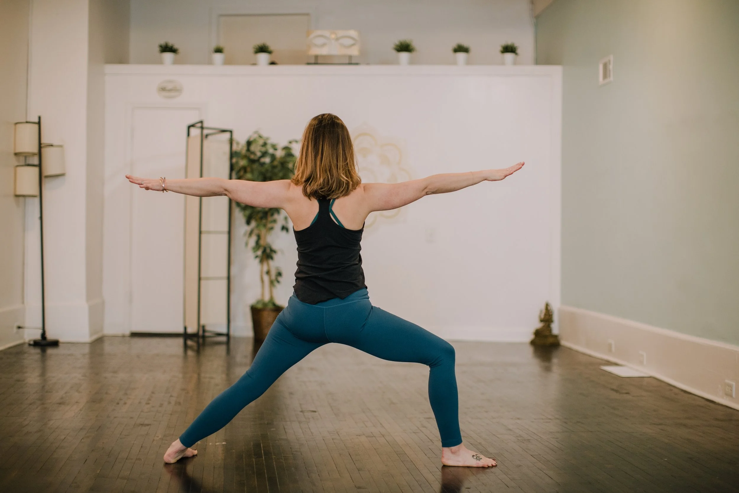 A woman practicing yoga in a studio, performing a Warrior II pose, with arms extended and feet firmly planted on the wooden floor.