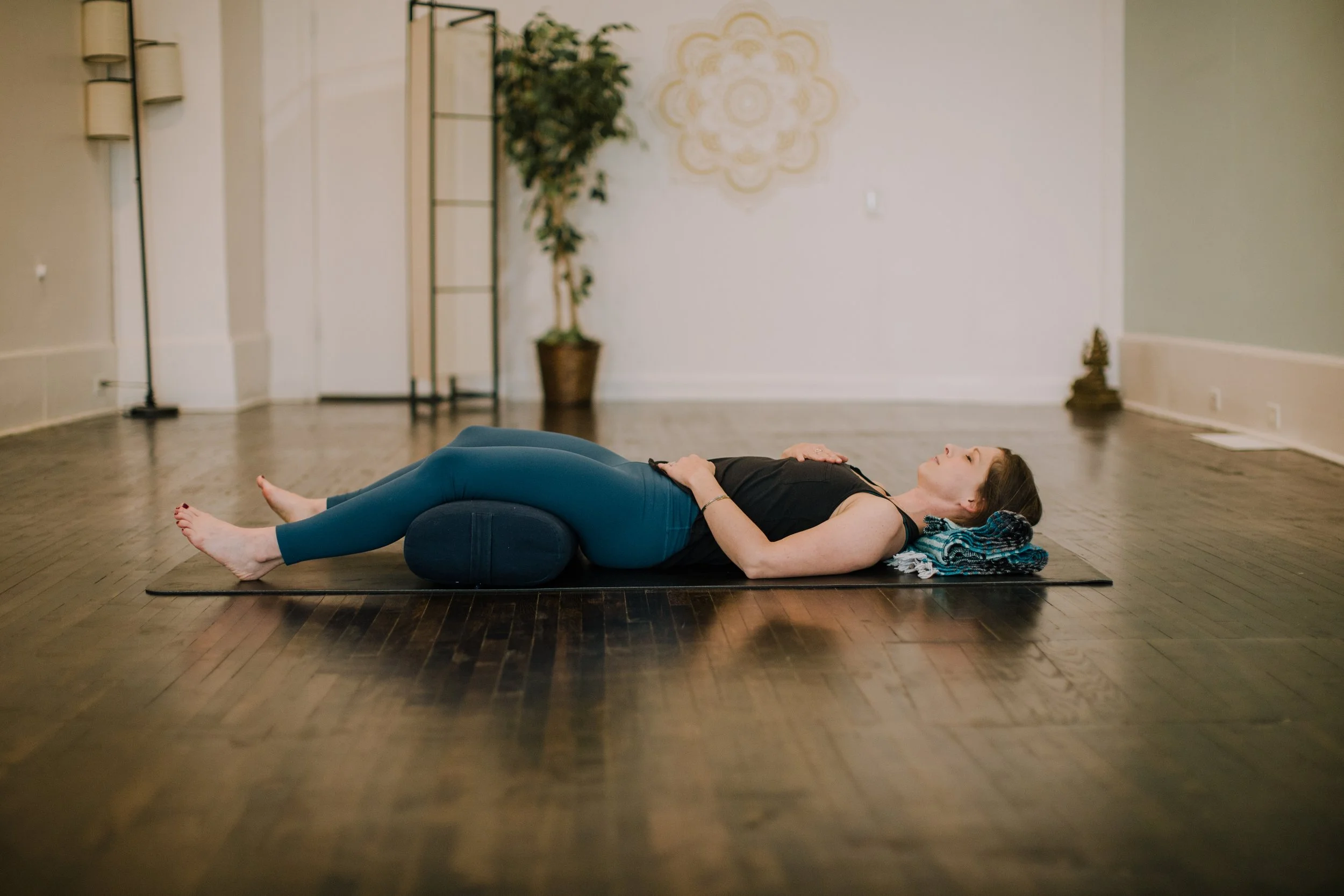 Person lying on a yoga mat in a relaxation pose with cushions, in a peaceful indoor setting with plants and soft lighting.