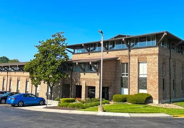 A two-story brick office building with modern architectural elements, surrounded by a landscaped area. Several cars are parked in front, under a clear blue sky.