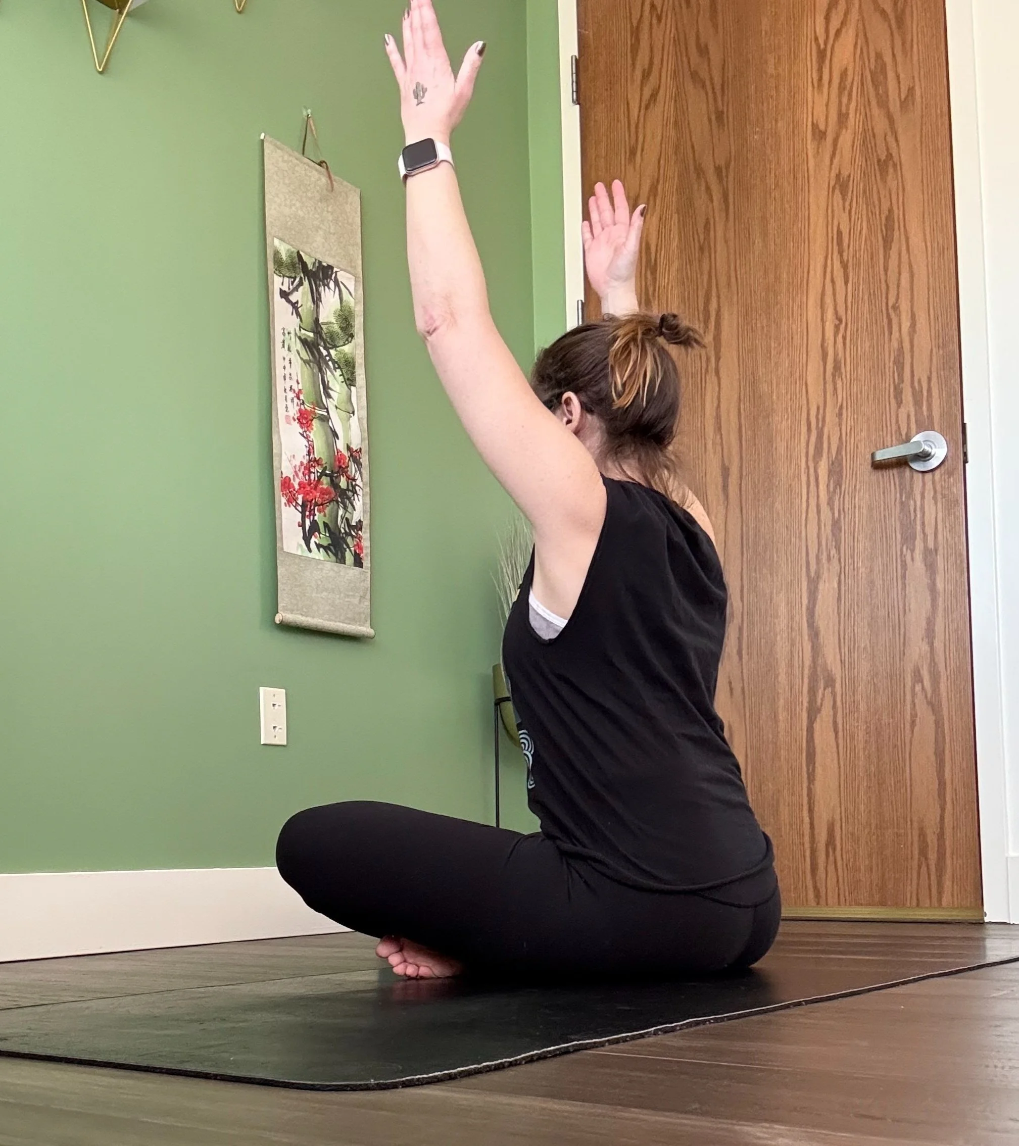 Person in black workout clothing practicing yoga with arms raised, sitting cross-legged on a mat in a room with green walls and a wall hanging. A wooden door is visible in the background.