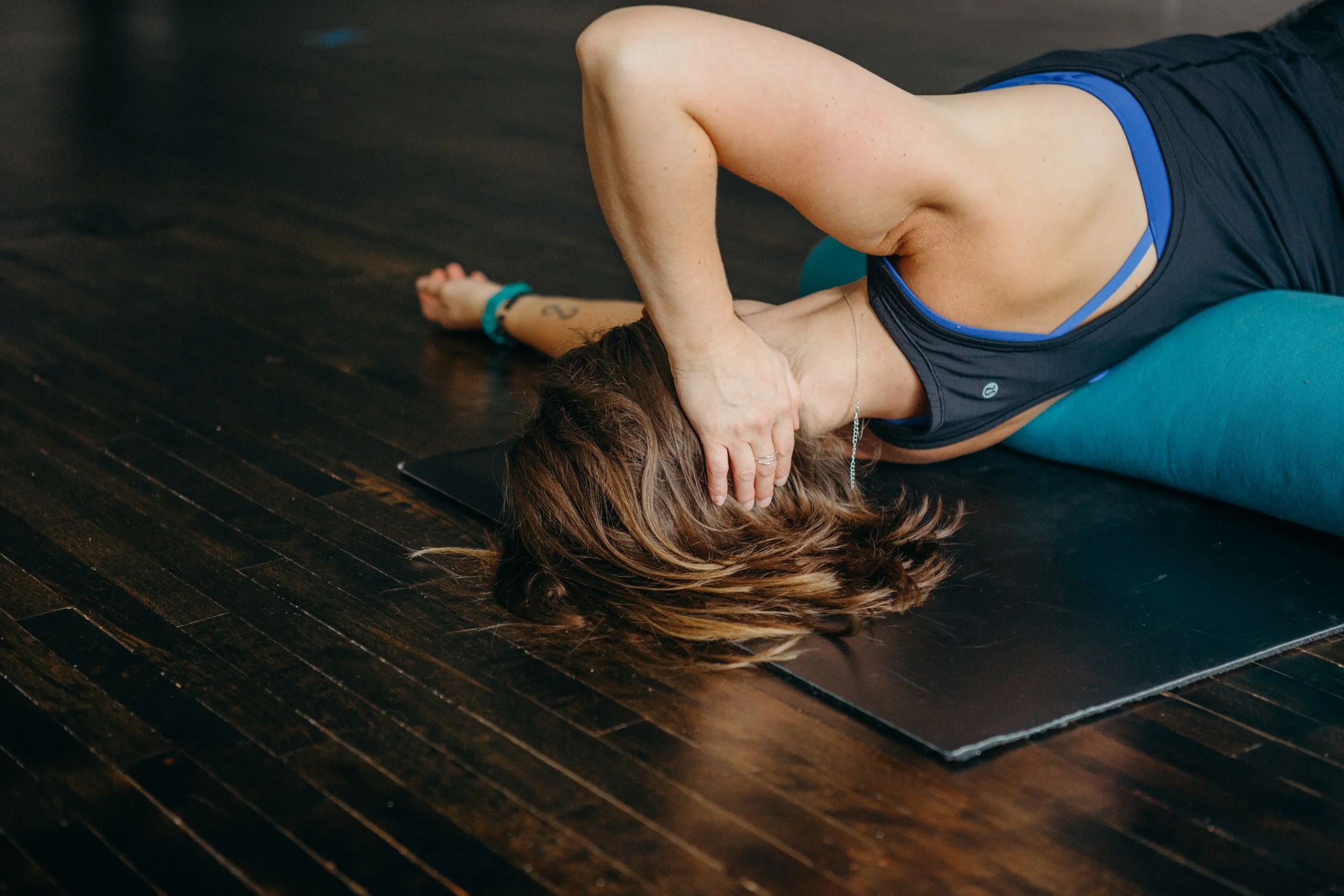 Person lying on floor doing yoga pose with arm overhead, resting on a mat and using a bolster for support, wearing athletic wear.