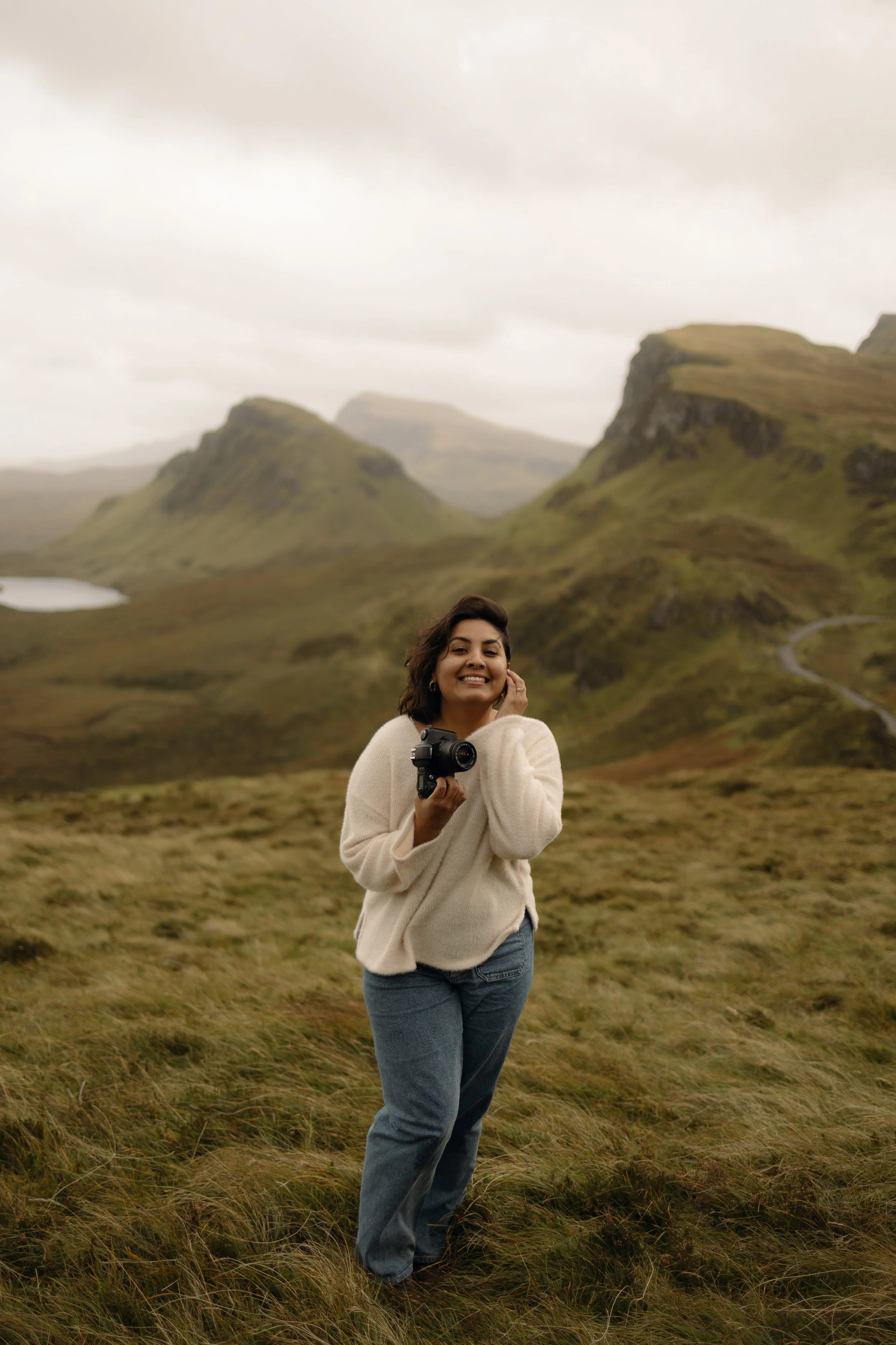Smiling woman holding a camera in a grassy mountain valley beneath misty cliffs