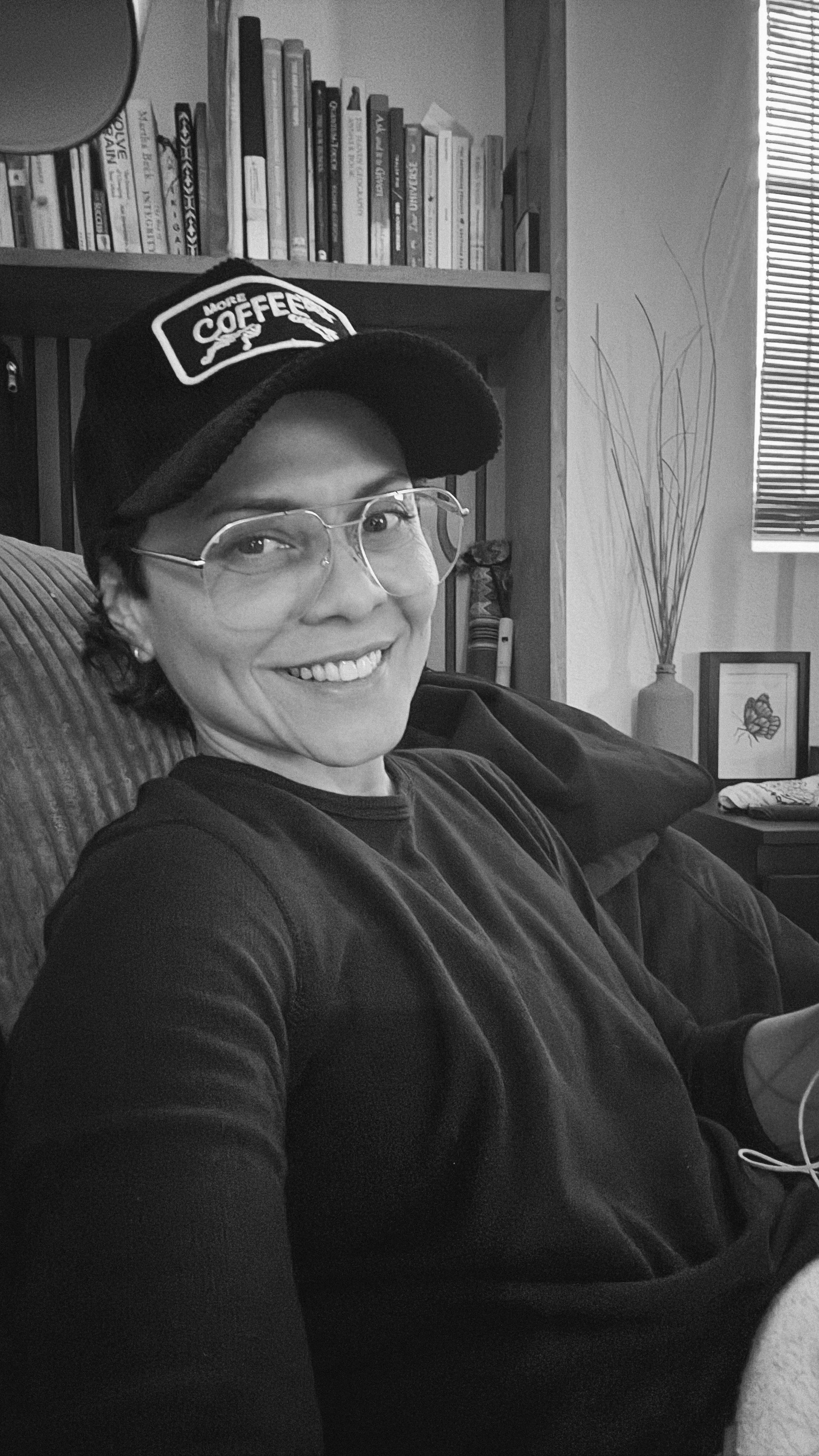 A woman with glasses and a cap smiling and sitting on a couch in a living room with a bookshelf, a framed butterfly picture, tall decorative branches in a vase, and a window with blinds in the background.