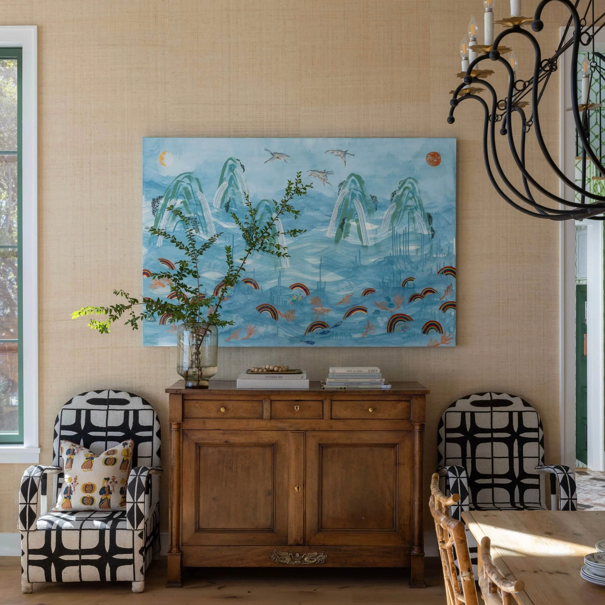 Interior of a room with a wooden sideboard, black and white patterned armchairs, a large colorful painting of mountains, water, rainbows, and birds on the wall, a glass vase with plant stems on the sideboard, and a black chandelier hanging from the c