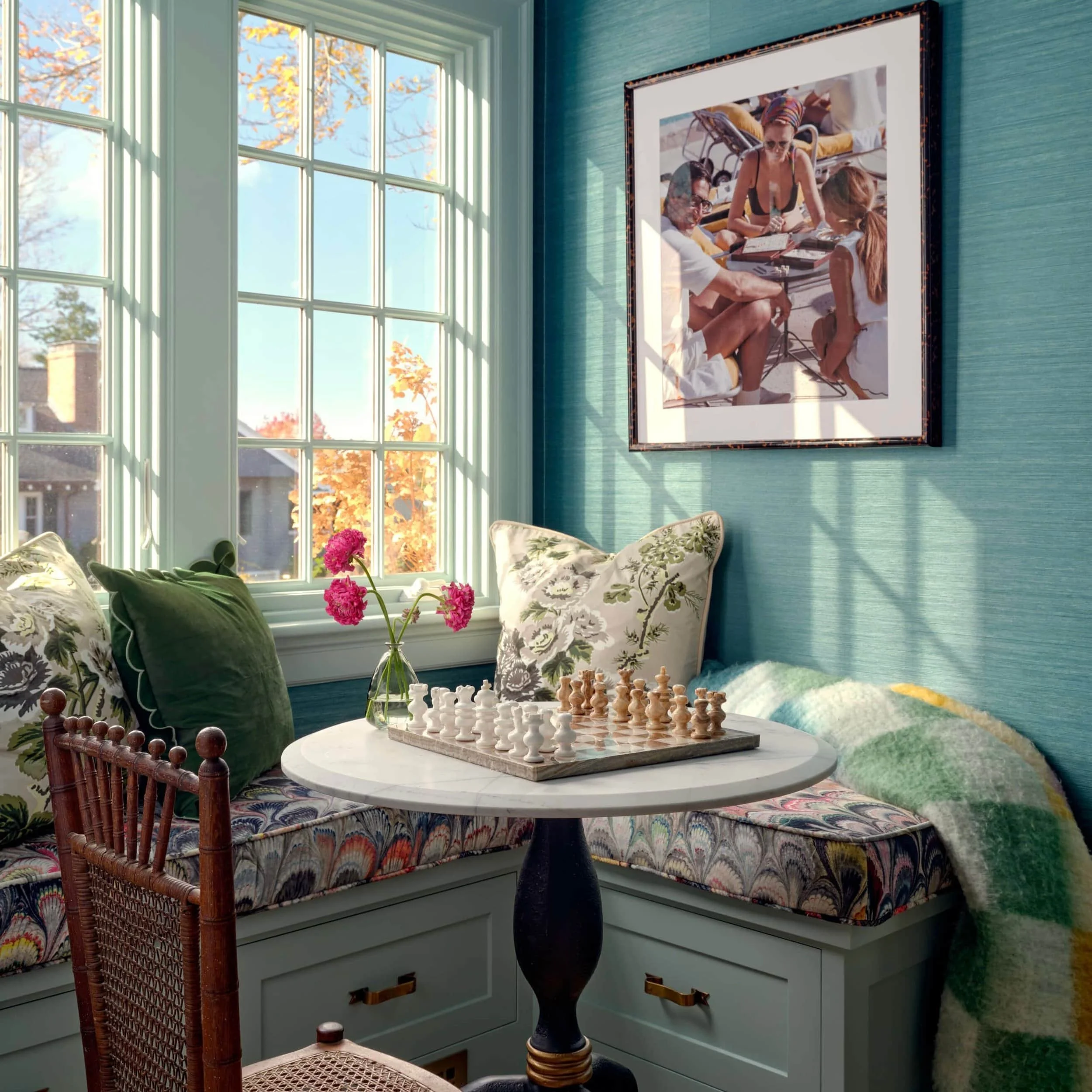 Sunlit breakfast nook with a window seat, floral cushions, a white round table with a chessboard, a small vase of pink flowers, and a blanket. A framed photo of a woman and children at the beach hangs on the teal wall.