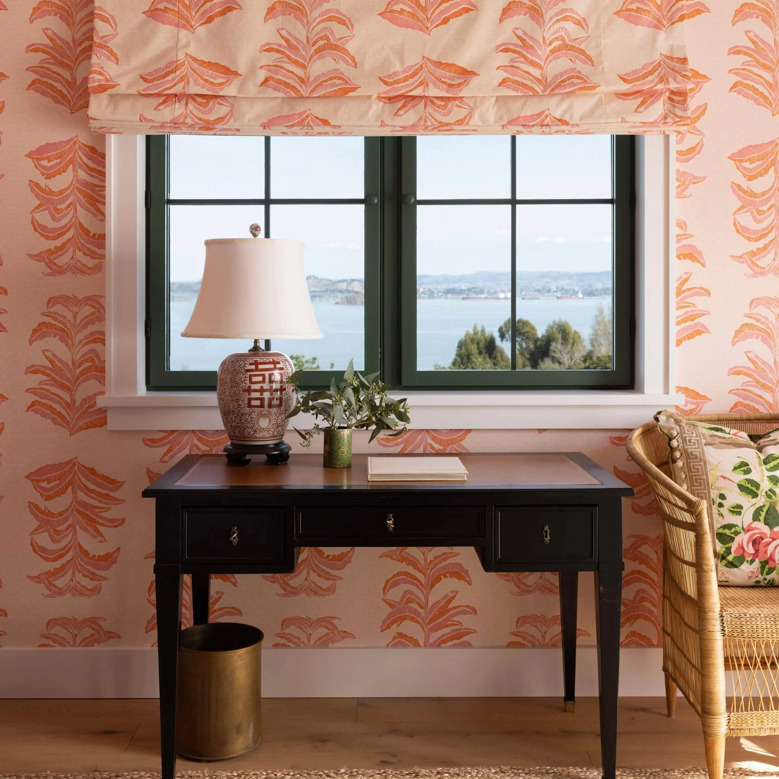 Interior room with a black desk, a ceramic table lamp, a small green plant in a vase, two books, and a window with a view of water and hills in the background, patterned wallpaper, and a rattan chair with floral cushion.