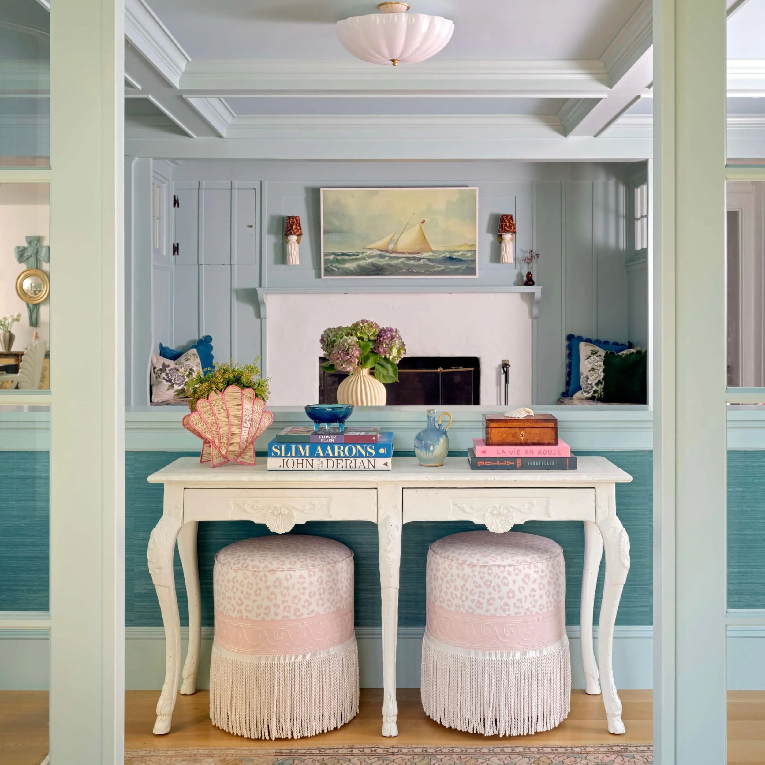 A cozy living room with a white vintage table, two pink upholstered stools with fringe, a vase of pink hydrangeas, and a decorative shell. The background has a fireplace, nautical paintings, and seating nook with pillows, in pastel-colored walls.