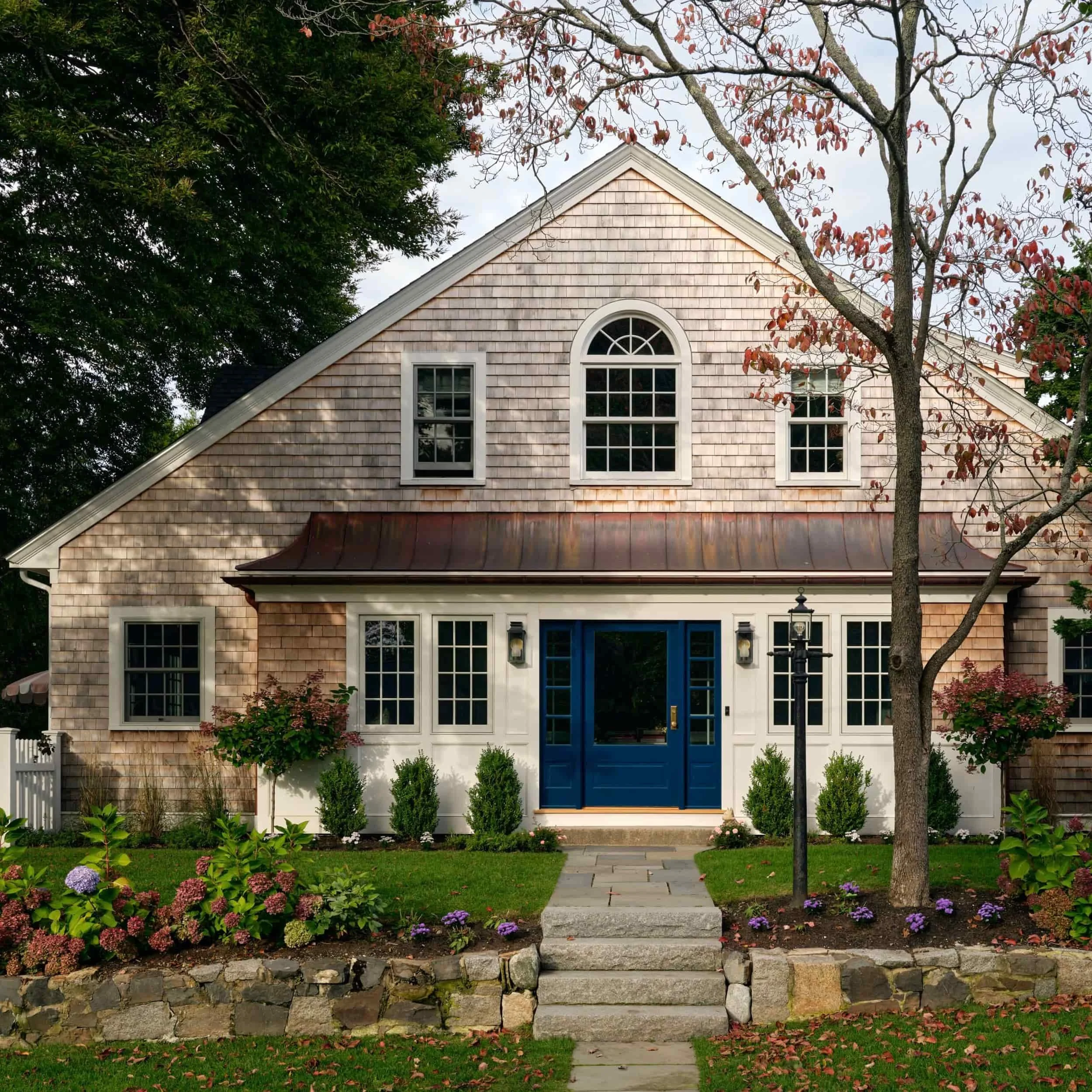 Front view of a house with a blue front door, brick and wooden shingle exterior, surrounded by a landscaped yard with trees, bushes, and flowers.