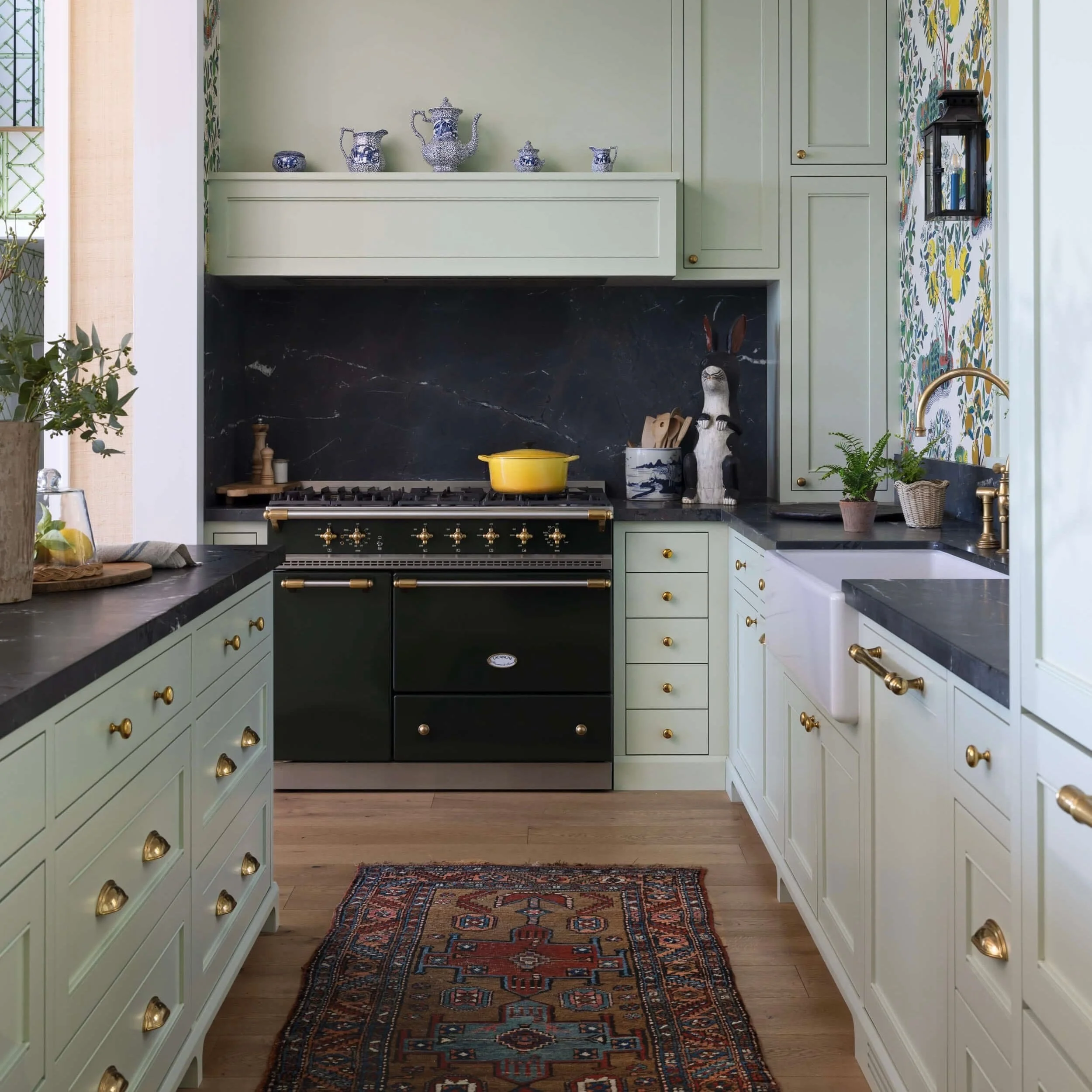 Kitchen with light green cabinetry, black countertops, and a black and gold stove. Tea pots on the upper shelf, decorative rabbit sculpture, potted plant, and a patterned rug on a wood floor.